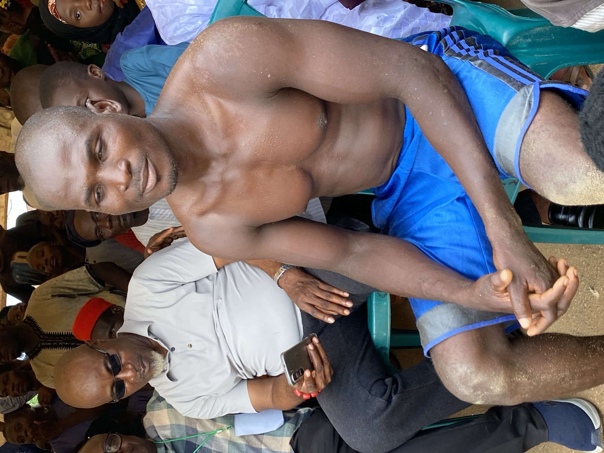 The image shows a group of people gathered in Kafanchan A, Nigeria. A shirtless man with a muscular build and blue athletic shorts is seated prominently. He is surrounded by other individuals, some of whom are also seated. One man wears sunglasses and a light-colored shirt, holding a phone. The overall environment appears to be outdoors, possibly a gathering of some sort. Many individuals are observed within this image.