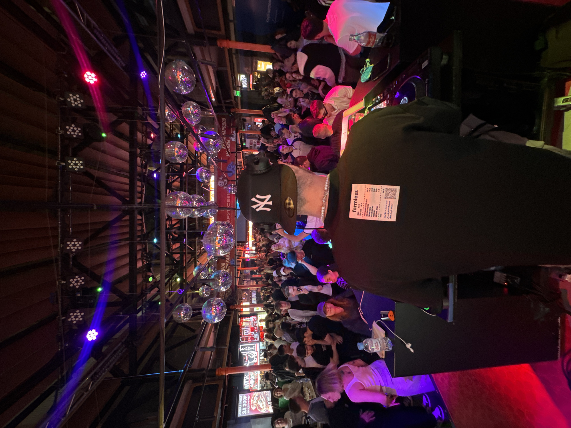 The image shows a crowded indoor space, possibly a club or venue, illuminated by disco balls and colorful stage lighting. There is a dense gathering of people in the background. In the foreground, an individual wearing a black jacket with a receipt or label attached to the back is visible. The person is also wearing a New York Yankees baseball cap. In the background, there are signs with Russian text and what appears to be a menu or advertisement for food, with prices displayed. The atmosphere suggests a lively or festive environment. It's likely that the scene is in Moscow, Russia, given the Cyrillic script on the signage in the background.