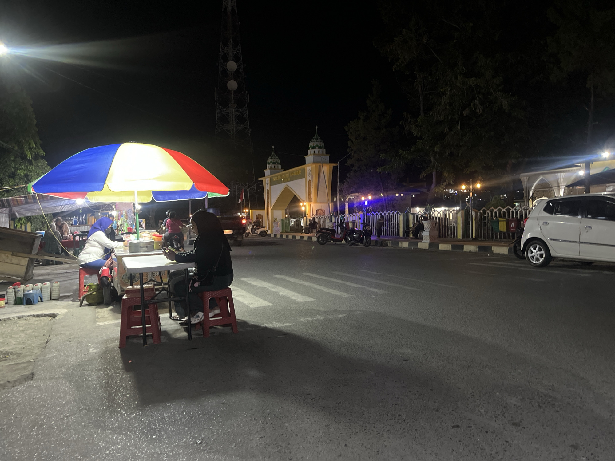 This image captures a nighttime scene on a street in Luwuk, Indonesia, likely near the Masjid Agung An-Nur. A brightly colored, multi-section umbrella, featuring red, yellow, blue, and green panels, is set up, providing shelter for a small food stall. Underneath the umbrella, two individuals are seated on red plastic stools at a white foldable table. One person, wearing a dark jacket and headscarf, is looking down at a mobile phone and appears to be holding it with both hands. Another person, wearing a white top and blue pants with a headscarf, is also seated at the table. Several plastic bottles and containers are visible on the ground near the stall, along with a yellow object that might be a bag or a mat.
Across the street, a white car is parked. In the background, a large transmission tower is visible, illuminated against the dark sky. Further down the street, an archway with a domed structure, characteristic of an Islamic architectural style, is prominent, with text that appears to read "Masjid Agung An-Nur Luwuk". Several motorcycles are parked near the archway. The street itself is paved and shows signs of wear, with a pedestrian crossing marked by white stripes. Trees line the side of the road, their branches silhouetted against the night sky. Streetlights and illumination from the stall and buildings create a dynamic interplay of light and shadow. The overall atmosphere suggests a lively, if modest, commercial area operating late into the evening.