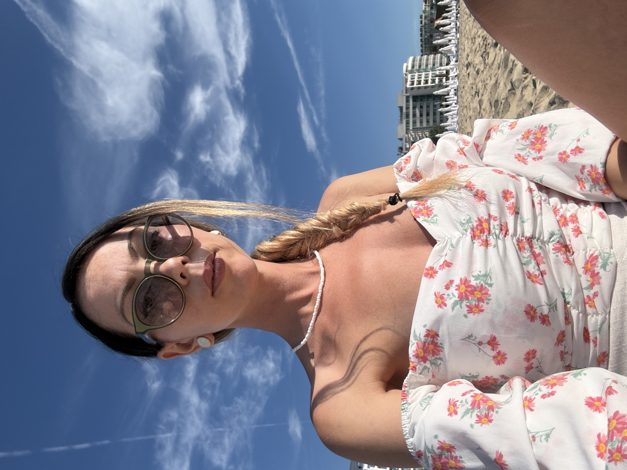 A woman is taking a selfie on a sandy beach in Nesebar, Bulgaria. She is wearing a white off-the-shoulder dress with a floral print, sunglasses, and a pearl necklace. Her blonde hair is styled in a braid. In the background, there are buildings and beach umbrellas. The sky is blue with white clouds. The sun is shining.