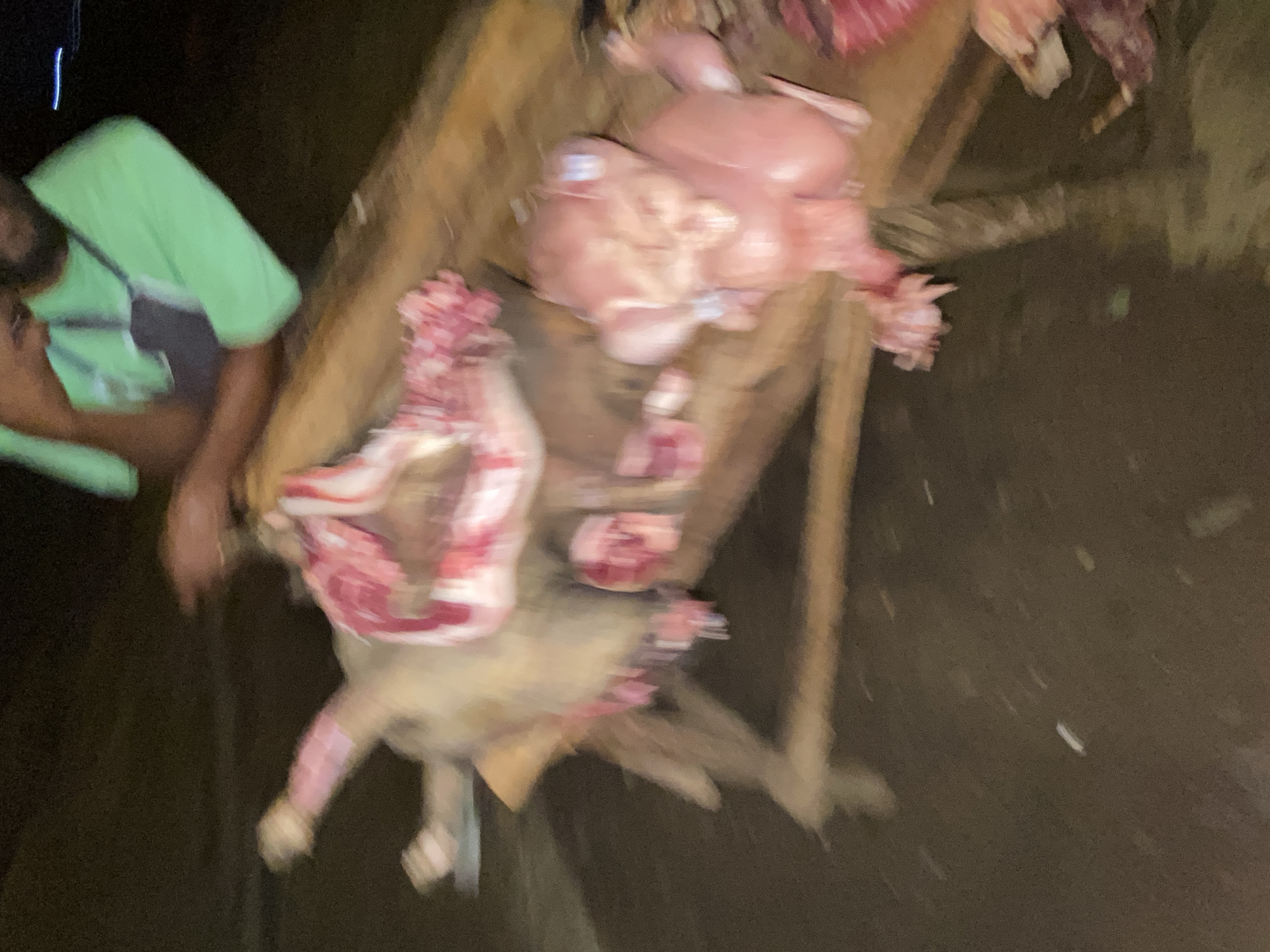 In a dimly lit environment in Bukuru, Nigeria, a person wearing a lime green shirt stands to the left of a makeshift wooden table. The table holds various cuts of meat, including what appears to be poultry and other unidentified pieces of raw flesh. The scene suggests a possible butchering or meat-selling operation. The ground around the table is dark and undefined.