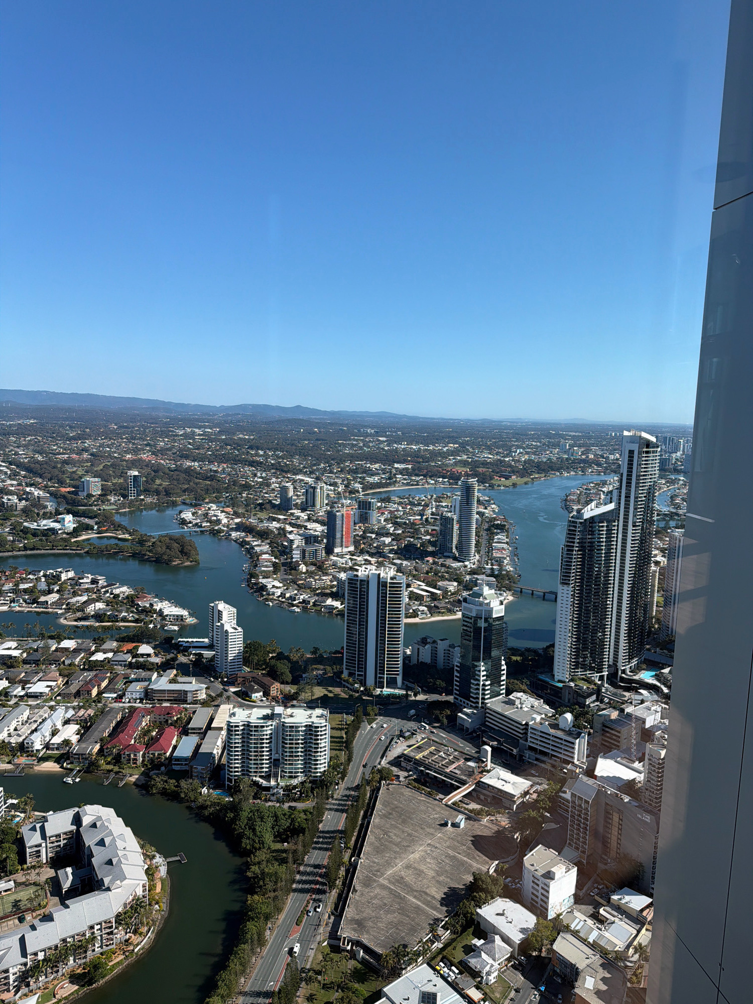 This is an aerial, wide-angle, high-angle view of the city of Gold Coast, Australia, specifically Surfers Paradise. The image captures a sprawling urban and waterway landscape under a clear blue sky.
In the foreground, a large modern building with a reflective facade, likely a skyscraper, dominates the right side of the frame, casting a shadow over parts of the scene.
The mid-ground features a wide, dark body of water, likely a river or canal, that snakes through the city. Along its banks are numerous residential buildings and apartment complexes, characterized by white and grey exteriors, many with balconies. Several moored boats are visible in the waterways.
Further into the cityscape, more high-rise buildings, including several distinct skyscrapers with modern architectural designs, are concentrated. One prominent skyscraper has a distinctive red accent on its upper floors. Roads with visible vehicle traffic crisscross the urban areas.
The background shows a dense suburban sprawl with houses featuring red-tiled roofs, interspersed with greenery. In the far distance, rolling hills or mountains form a hazy horizon line. The overall scene depicts a sun-drenched, developed coastal city with extensive waterways and a mix of residential and commercial architecture.