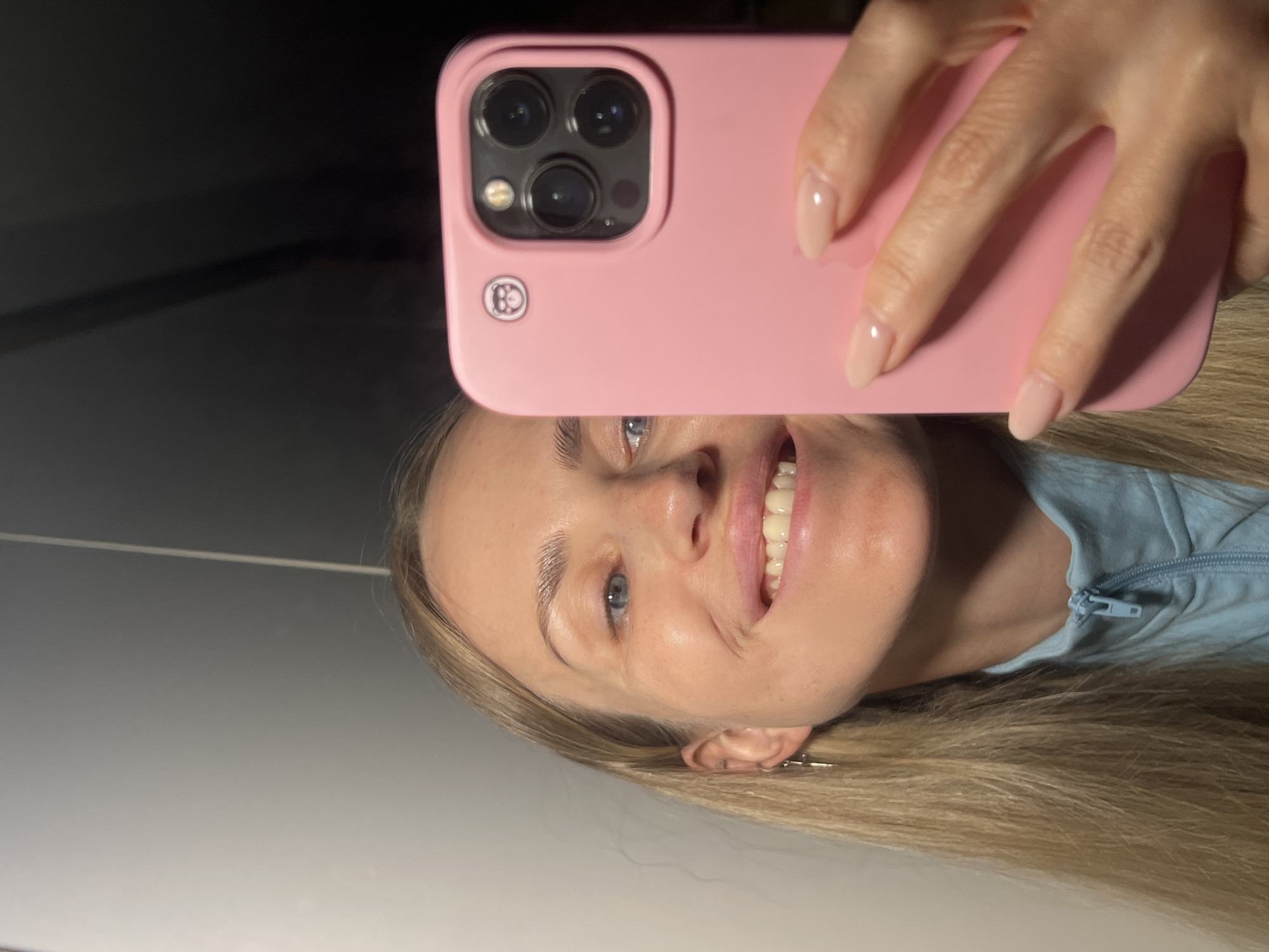 A woman is taking a selfie with her iPhone. She is holding the phone in her right hand, and the back of the phone is visible. The phone has a pink case with a camera bump. The woman has blonde hair and is wearing a light blue zip-up jacket. Her nails are painted a light pink color. The background is out of focus and appears to be a wall with some dark shadowing. The scene is indoors.