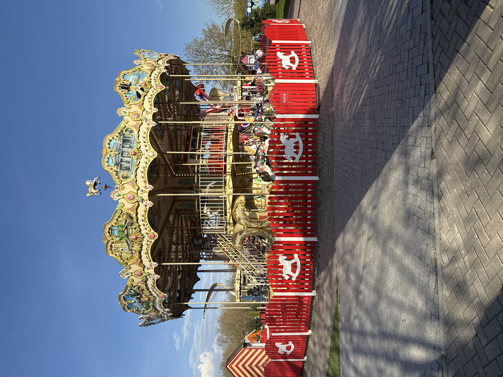 The image depicts a carousel in Khimki, Russia, under a clear blue sky. The carousel is enclosed by a red fence adorned with white rocking horse figures. The surrounding area includes a brick walkway and some greenery, suggesting a park setting.
