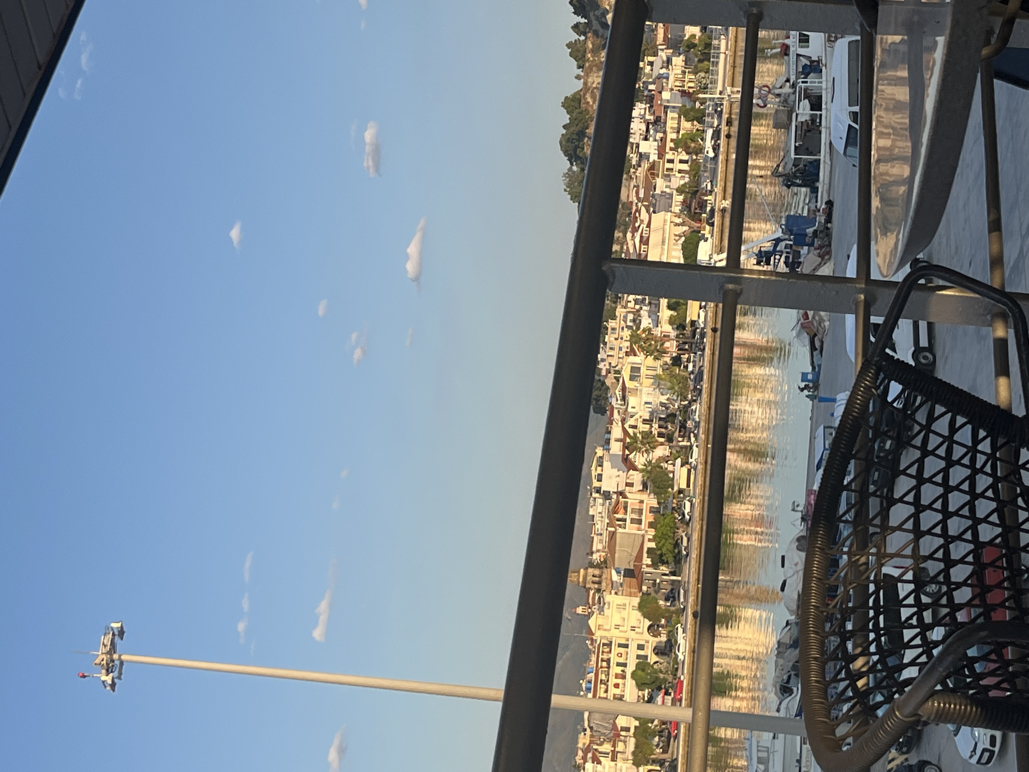 This is a telephoto, eye-level, outdoor shot captured during daylight, showcasing a coastal town with a harbor. The foreground features the top of a dark woven chair and a metal railing, partially obscuring the view. Beyond the railing, a body of water reflects the warm hues of the buildings lining the waterfront. A cityscape with multi-story buildings, featuring terracotta roofs and light-colored facades, stretches along the coast. Trees are visible interspersed among the buildings and on a hill in the background. The sky is a clear blue with a few scattered white clouds. A tall lamppost stands to the left of the frame, with multiple lights and antennas at its peak. Several boats are docked in the harbor, including a white ferry or passenger vessel and smaller fishing boats. A few vehicles are visible on a road or promenade in front of the buildings. The overall impression is of a peaceful harborside scene in a Mediterranean town.