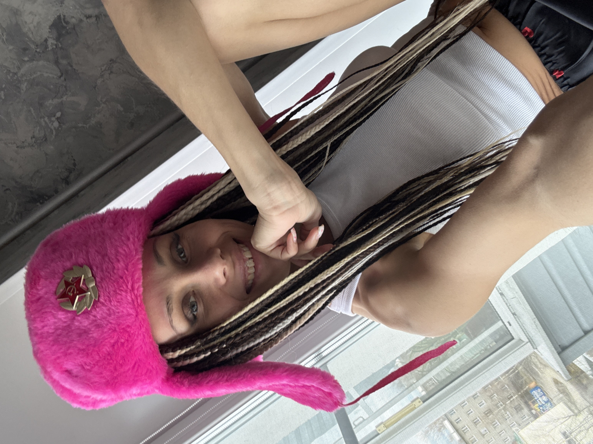 A woman with blonde and brown braids, wearing a bright pink ushanka hat with a Soviet Union emblem, is smiling and posing for a selfie in front of a window. She is wearing a white tank top and has her hand near her face. Outside the window, a cityscape is visible, suggesting an urban location. The specific location of Yaroslavl', Russia is not discernible without additional information.