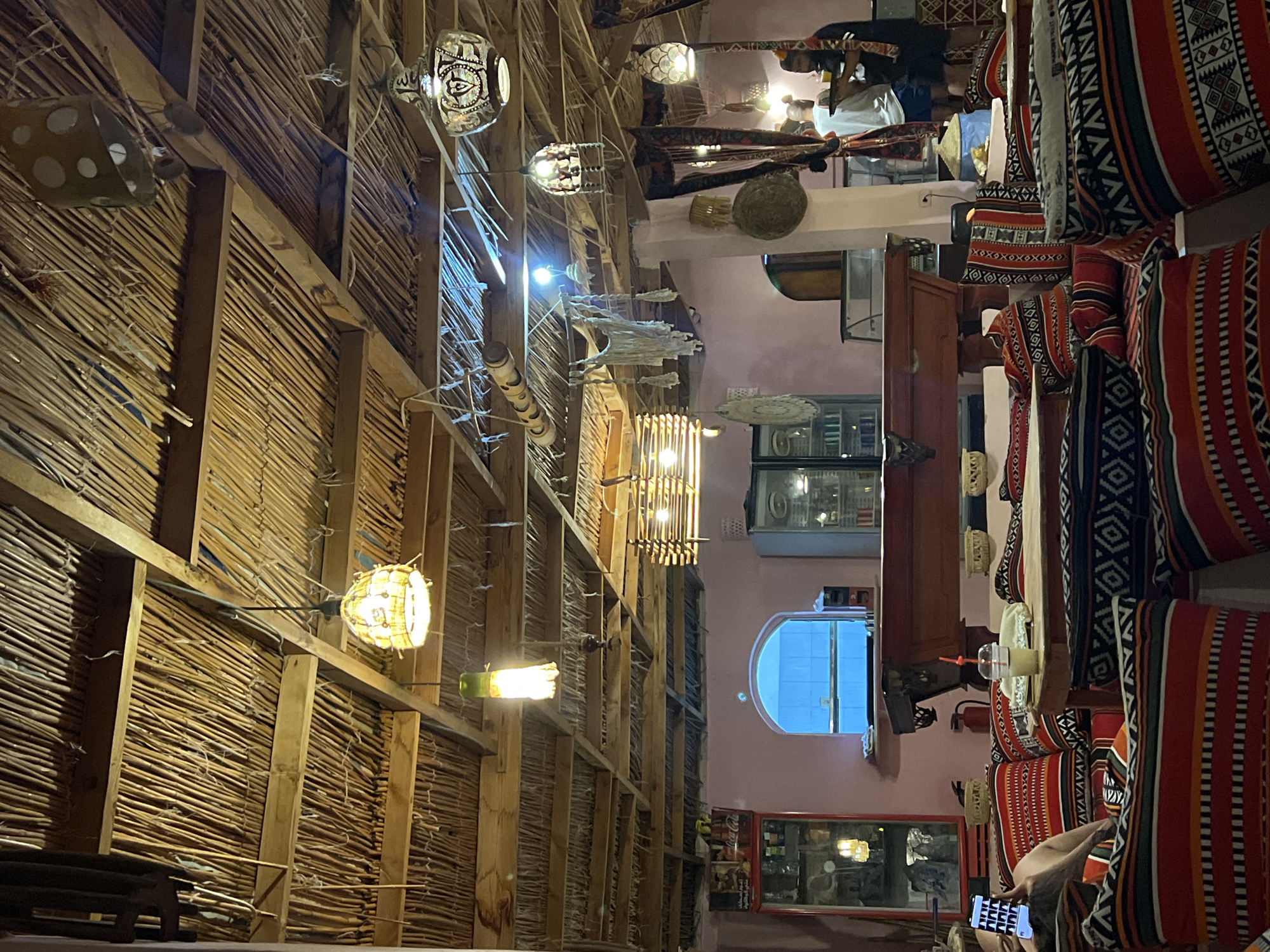 The image depicts the interior of a cafe with a distinct Bedouin-inspired aesthetic, likely located in Nueiba, Egypt. The ceiling is constructed from natural materials, possibly palm fronds or reeds, supported by wooden beams, and adorned with numerous hanging lamps of various designs, including some with intricate metalwork and others with a more rustic, textured appearance. The walls are painted a soft pink, and an arched window with a bright blue pane offers a view to the outside.
The seating area is characterized by low, floor-level tables and an abundance of large, patterned cushions in vibrant colors, predominantly red, black, and white, with geometric and tribal motifs. These cushions are arranged to create a comfortable lounge-like atmosphere. In the foreground, a person is visible sitting on these cushions, holding a smartphone.
Further into the cafe, a large, wooden table, possibly a pool table, is centrally positioned. Behind it, there are refrigerators and display cases, suggesting a food and beverage service. Two men are visible in the background, one appearing to hold a tray. The overall environment is warm and inviting, with soft lighting from the lamps contributing to a cozy ambiance. The decor strongly suggests a cultural theme, common in tourist areas of Egypt, particularly those with a Bedouin heritage.