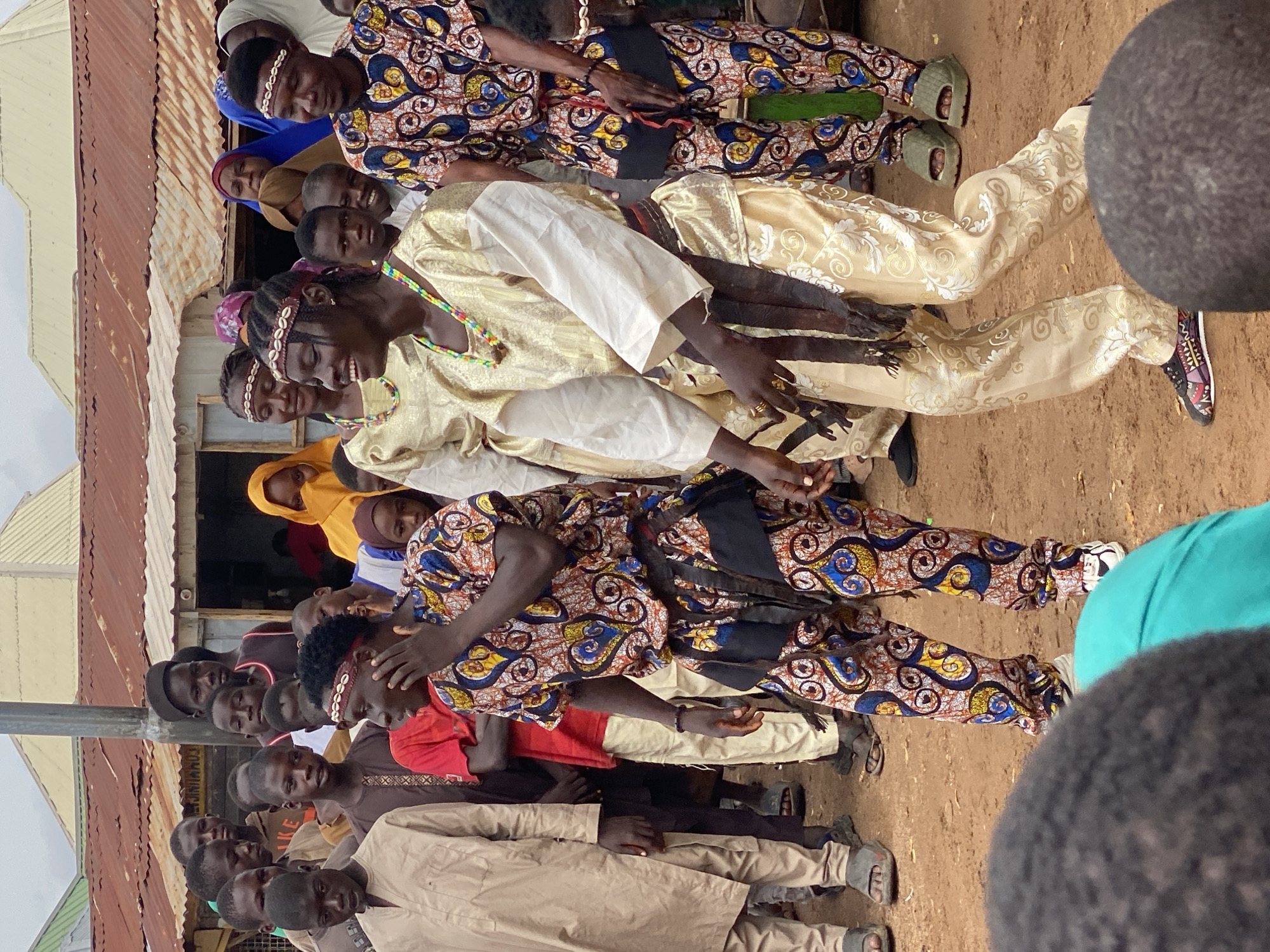 The image shows a group of people in what appears to be an outdoor gathering in Rigachikun, Nigeria. The subjects are predominantly young men and boys, with some women and girls also visible. They are dressed in a mix of traditional and modern clothing. Several individuals are wearing vibrant, patterned fabrics, including anoraks and matching trousers. Some are also adorned with headbands made of shells or beads. One person in the foreground is wearing a golden-colored robe and a beaded necklace.
The scene seems to be set in a somewhat informal environment, possibly a village or community space, with corrugated metal roofing and dirt ground visible. The lighting suggests it is daytime, though the weather is overcast. The people are positioned in a way that suggests they are either watching something or participating in an event. Some are looking towards the center of the group, while others are looking towards the camera. Their expressions are varied, with some appearing engaged and others more subdued. The overall mood seems to be one of communal gathering, possibly a celebration or a social event. There is no visible text in the image.