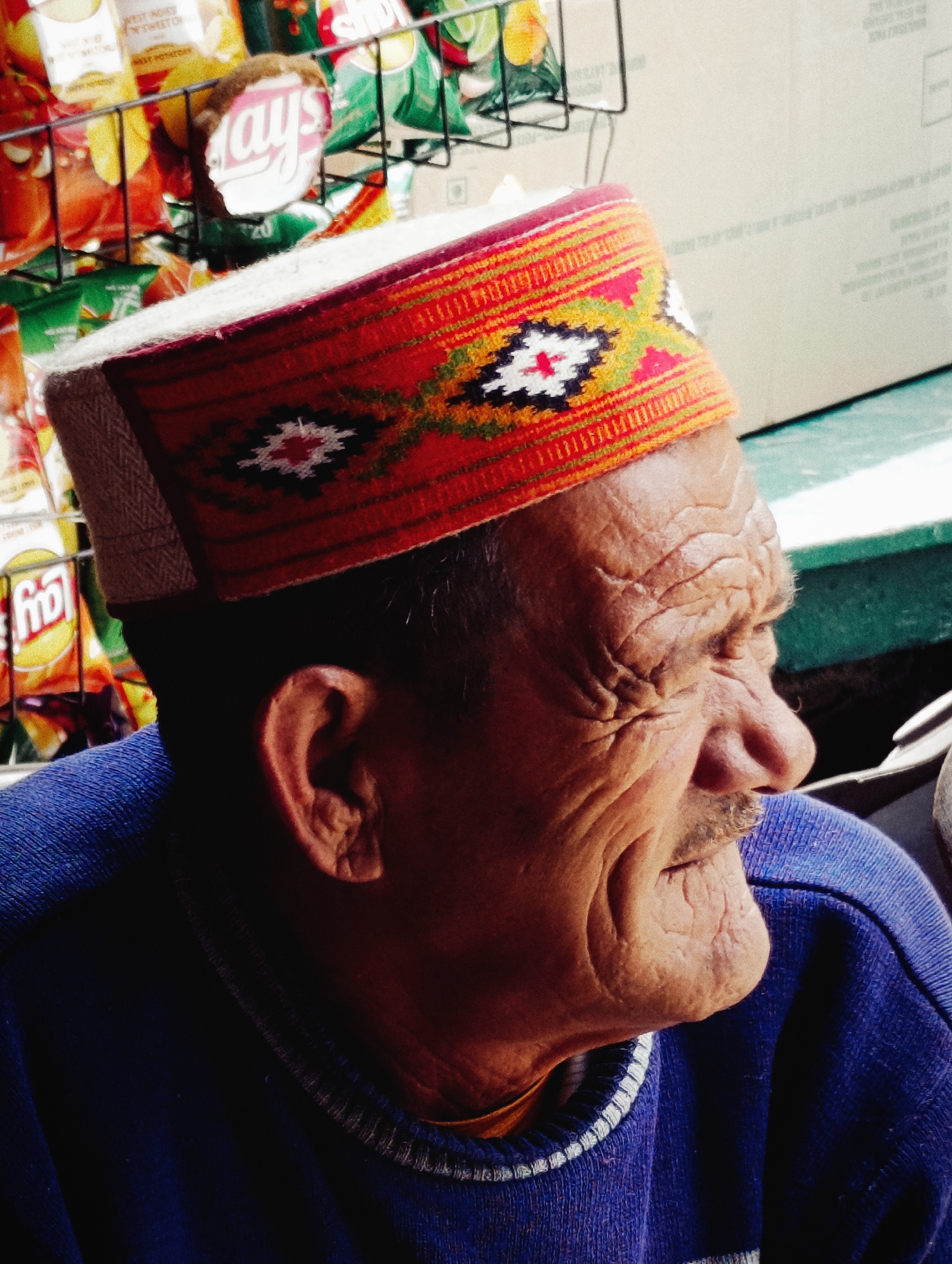 An elderly man with deeply wrinkled skin and a grey mustache is wearing a traditional Himachali cap with red, orange, yellow, and white geometric patterns. He is also wearing a blue sweater. The man is positioned in profile, looking towards the right side of the frame. In the background, a rack of snack bags is visible, with a prominent "Lays" bag. A large, light-colored cardboard box is also present in the background, suggesting an outdoor market or roadside stall. The scene appears to be set in Kothi, India, a popular tourist destination in Himachal Pradesh known for its scenic beauty and local crafts.