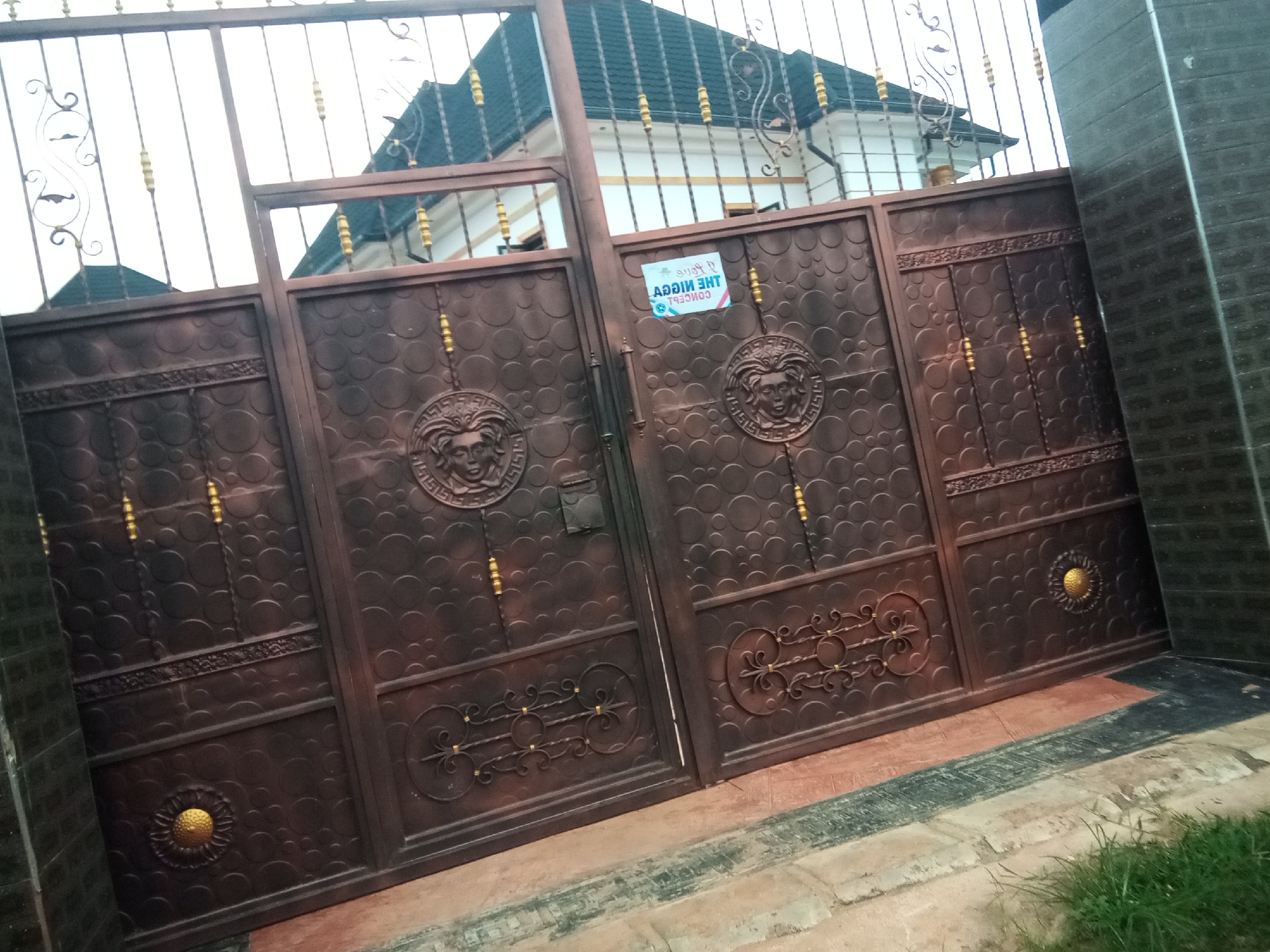 The image shows a large, ornate metal gate with a dark brown finish and decorative gold accents. The gate has two main panels, each featuring a raised circular design with a lion's face. There are also smaller gold accents and intricate scrollwork on the gate. A small, rectangular sticker is attached to the right panel of the gate, displaying text that is partially obscured and reversed. Behind the gate, a white building with a dark, pitched roof and decorative white railings is visible. The building appears to be a house. The gate is set against a backdrop of a cloudy sky. In the foreground, there is a paved walkway with reddish-brown and black sections, and a strip of green grass. The overall scene suggests a residential property. The image is geotagged to Nanka, Nigeria.