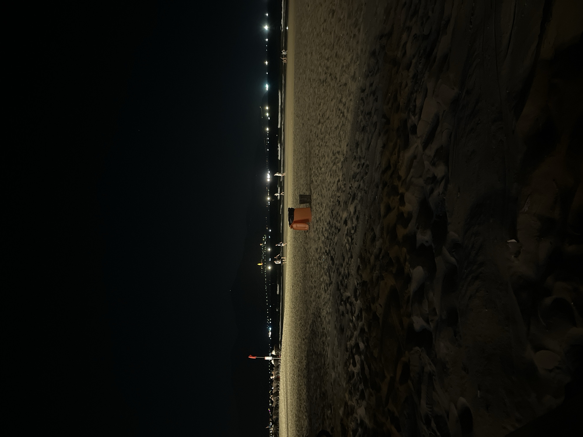 The image depicts a nighttime scene at a beach. The foreground consists of dark sand, illuminated slightly, revealing its texture. In the middle ground, there is a lifeguard stand and other people present on the beach. Further into the distance, a row of lights suggests the presence of a city or town along the coastline. The left side of the image is dominated by a dark sky. The location is likely a beach in Đà Nẵng, Vietnam, given the coastal setting and urban development. The people are interacting with the beach environment.