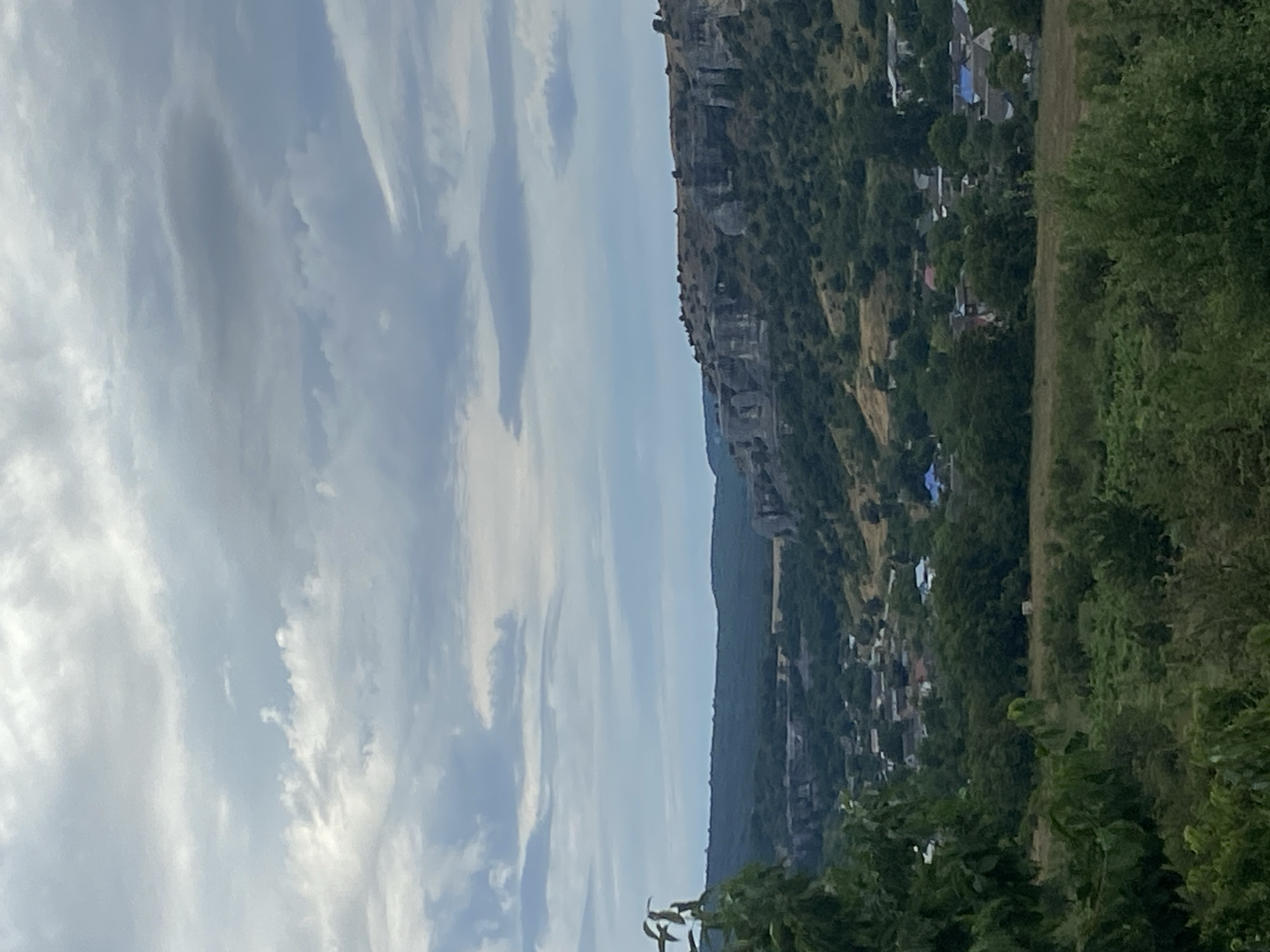 The image depicts a rural landscape with rolling hills and a distant cliff face. In the foreground, lush green foliage is visible. A small village with houses and blue roofs is nestled within the valley. The sky is overcast with scattered clouds. The scene appears to be a serene, undeveloped natural environment with a small settlement integrated into the landscape.