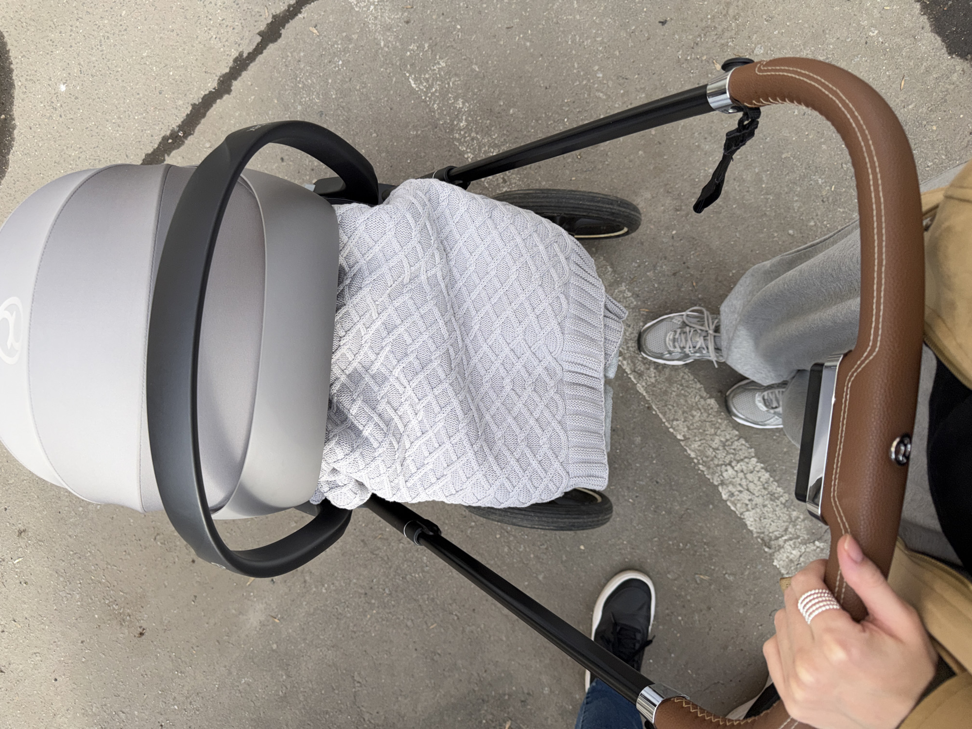 The image presents an overhead view of a modern baby stroller being pushed or attended to outdoors on a paved surface. The primary subject is a light grey stroller with a sleek design, featuring a dark grey frame and black wheels. A light grey knitted blanket with a distinct diamond pattern is draped inside the stroller's bassinet or seat, suggesting a baby is present, though not visible.
A person is partially visible in the bottom right, holding the curved handlebar of the stroller. The handlebar is wrapped in brown leather with contrasting white stitching. The person's right hand, adorned with a silver multi-band ring on the index finger, grips the handle. Part of a tan or brown coat/jacket is also visible on this person, along with their left foot wearing a black sneaker near the bottom left of the frame.
Another pair of feet, belonging to a different person, are visible near the front wheel of the stroller. These feet wear grey athletic shoes and grey sweatpants. The ground is a grey, textured paved surface, consistent with concrete or asphalt, showing some minor wear and stains. A partial white painted line runs across the ground in the mid-right section of the image.
A subtle white logo, appearing to be a stylized "C" or "Cy" with a circular element, is visible on the side of the grey stroller body. The lighting is bright and even, indicating it is daytime. The scene captures an ordinary outdoor activity, likely a stroll, set in an urban or suburban environment within Khimki, Russia. No specific emotions are discernible.