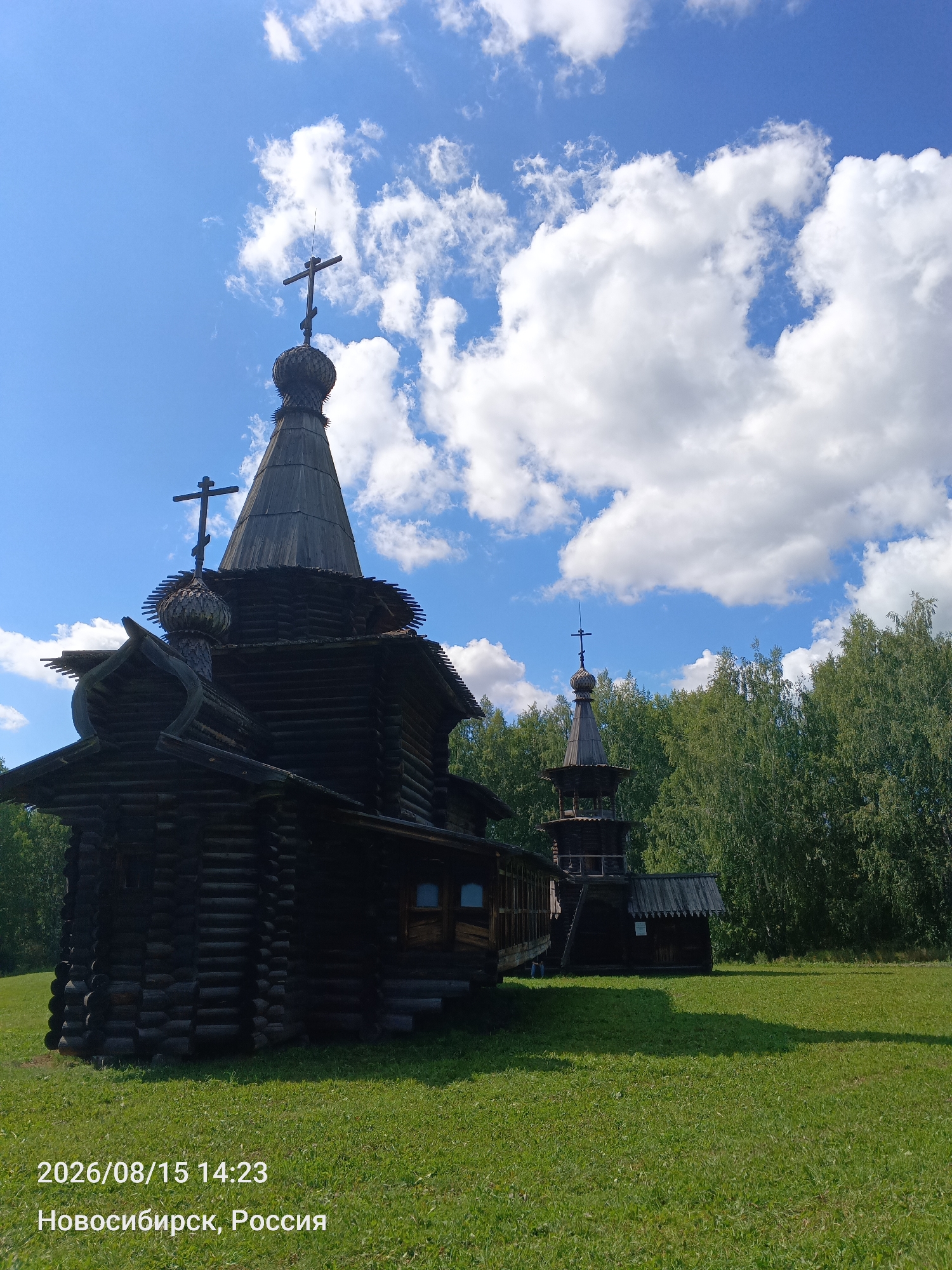The image shows a traditional wooden Orthodox church and a bell tower situated on a grassy field, with a dense forest in the background under a blue sky with scattered white clouds. The architecture of the church is dark wood, with multiple tiers and onion domes topped with crosses. The bell tower is also constructed of wood and has a similar architectural style. The scene is bathed in sunlight, casting shadows on the grass. A timestamp and location are visible in the bottom left corner: "2026/08/15 14:23" and "Новосибирск, Россия" (Novosibirsk, Russia). No people are visible in the image.