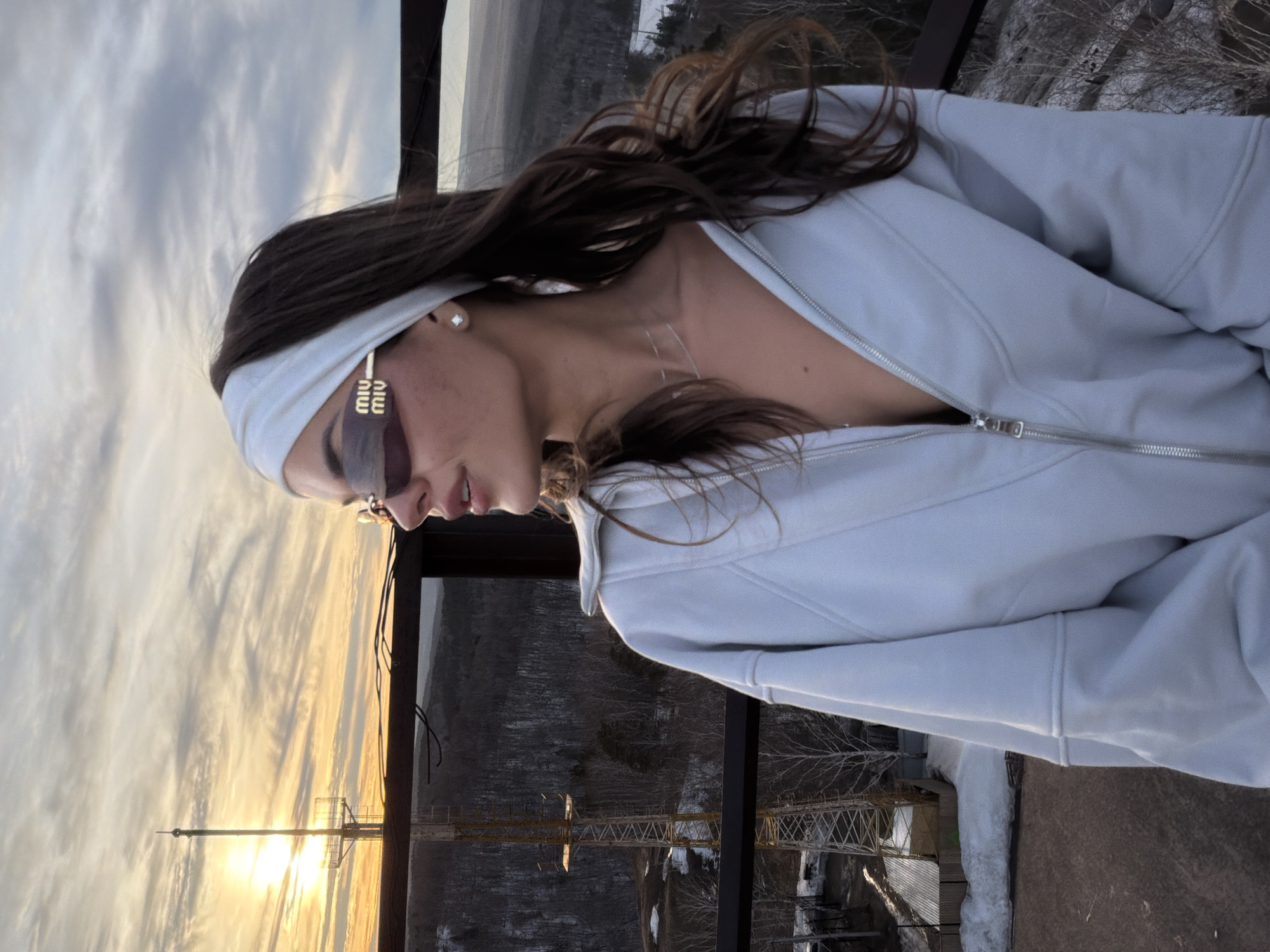 A woman stands on a metal platform, likely an observation deck or structure, overlooking a landscape near the city of Krasnoyarsk, Russia. She wears a light-colored zip-up jacket, a white headband, and sunglasses with the brand "Miu Miu" visible on the side. She also sports a small earring. Her dark hair is styled and falls over her shoulder.
In the background, a sunset is partially visible behind a metal communications tower. The landscape below includes a forest, structures, and possibly some areas with snow. The platform she is on is constructed of metal with a railing, and has traces of snow or ice.