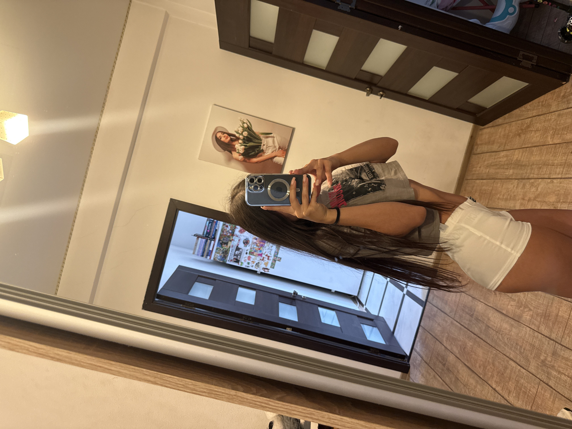 A young woman with long, dark hair is taking a mirror selfie in what appears to be a bedroom or hallway. She is wearing a grey crop top and white shorts. She is holding a smartphone in her right hand and is angled towards the mirror. In the reflection, another door is visible with several square glass panes. A framed picture of a woman holding flowers hangs on the wall to the right of the second door. The floor is made of wooden planks. The room has neutral-colored walls and ceiling with a light fixture.