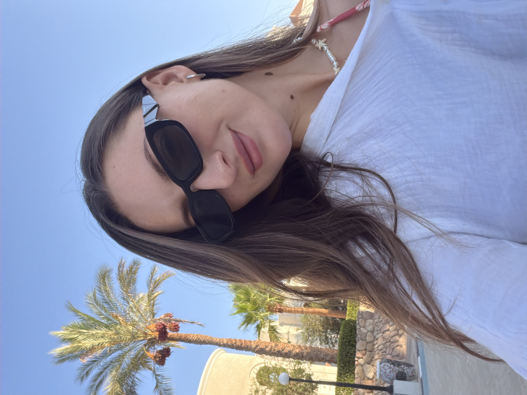 A young woman with long, dark brown hair, wearing black sunglasses, a white shirt, and a beaded necklace, smiles at the camera. Behind her, a tall palm tree with dates is visible against a clear blue sky. In the background, there are palm trees, a building with arched windows, stone walls, and a lamppost. The scene appears to be in Sharm El Sheikh, Egypt, suggesting a warm, sunny day.