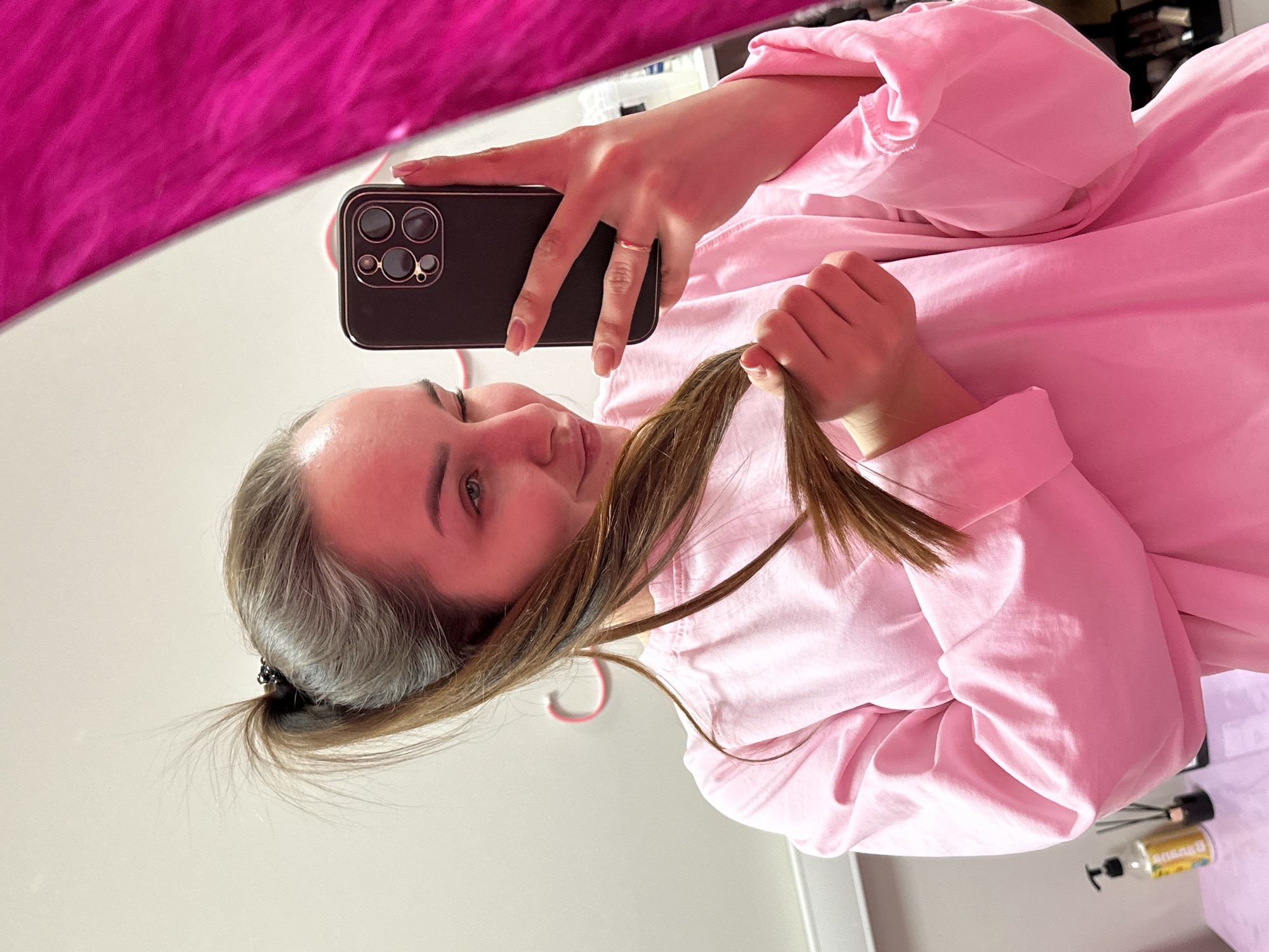 A woman is taking a selfie with a smartphone. She is wearing a pink sweatshirt and holding a strand of brown hair in her right hand. She has a ring on her left hand. Her hair is pulled back in a ponytail. The background is a mix of pink and white. A pink hanger is partially visible. There is a bottle of liquid and some other objects on a surface behind her. The location is likely indoors, possibly in a home. There is no definitive evidence in the image to indicate that it was taken in Chaikovsky, Russia.