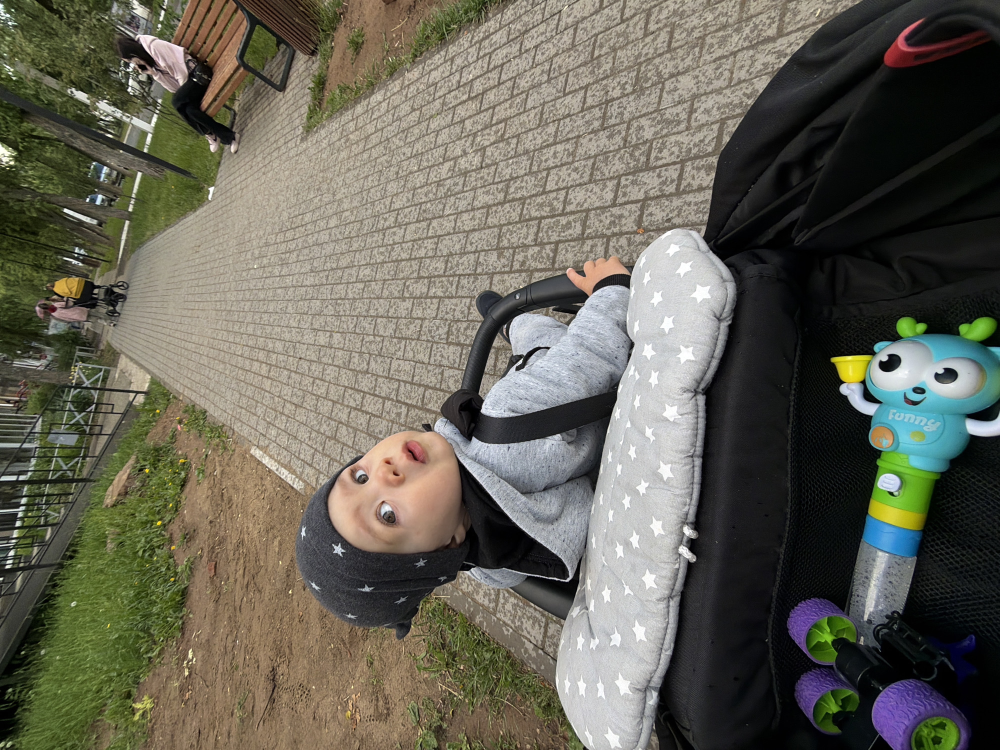The image, taken in Chaikovsky, Russia, captures a close-up, tilted view of a baby in a stroller on an outdoor paved path during the daytime. The baby, likely a toddler, is wearing a grey hooded top and a dark grey hat adorned with white stars. They are securely strapped into the stroller, which features a grey insert with white star patterns. The baby looks directly at the camera with wide, curious eyes and a slightly open mouth.
To the right of the baby, nestled within the stroller's compartment, are several toys. A prominent blue, green, and yellow toy resembling a character with "funny" written on its body is visible, along with a multi-colored base that suggests it might be a bubble wand. Below this, a purple and green wheeled toy is also present.
The stroller is positioned on a grey brick pathway. In the background, further down the path, a person wearing a pink top and dark pants is sitting on a wooden park bench. Beyond them, another person can be seen pushing a stroller. The path is lined with green grass and trees, with patches of dirt visible. The overall lighting suggests a mild, possibly overcast, day.