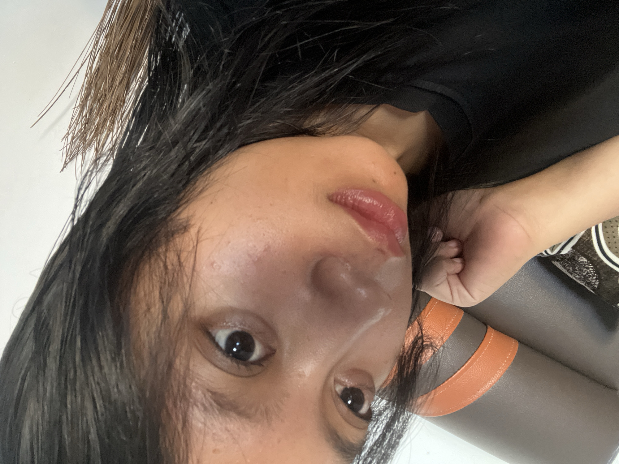 A close-up, angled, selfie-style photograph shows a young woman's face, primarily focusing on her eyes, nose, and mouth. Her dark hair falls across her face, partially obscuring her forehead and left eye. She appears to be wearing a black t-shirt. The background is out of focus but suggests an indoor setting with a white wall on the left and a brown and orange textured surface, possibly upholstery, on the left side of the frame. The overall lighting is soft, highlighting the sheen on her skin and the gloss on her lips. The image does not provide any information about the specific location within Jambearum, Indonesia.