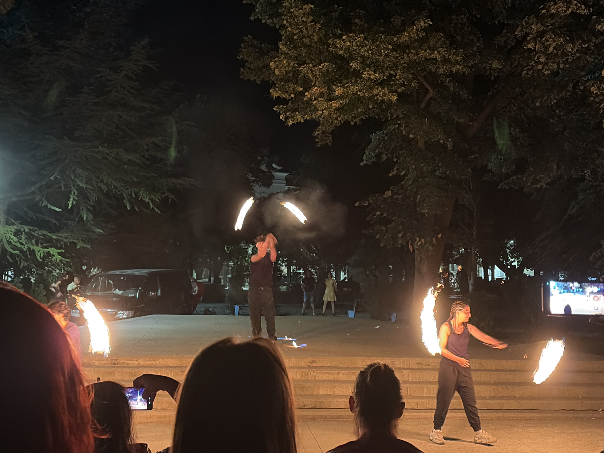 The image depicts an outdoor nighttime event in the city of Vratsa, Bulgaria, possibly a street performance or festival. In the foreground, several audience members are visible, with their backs to the camera, suggesting they are watching a performance. A smartphone is being used to record the event. The central focus of the image is a fire performance, with two individuals actively engaged in manipulating flaming objects. One performer, a male, is in the mid-ground, appearing to spin fiery props that create arcs of light and smoke. Another performer, a female, is to the right, also with flaming props. The background shows trees, buildings, and a dark sky. The ground appears to be paved, with some raised steps or a stage area. The presence of fire and the crowd indicates a lively, public gathering.