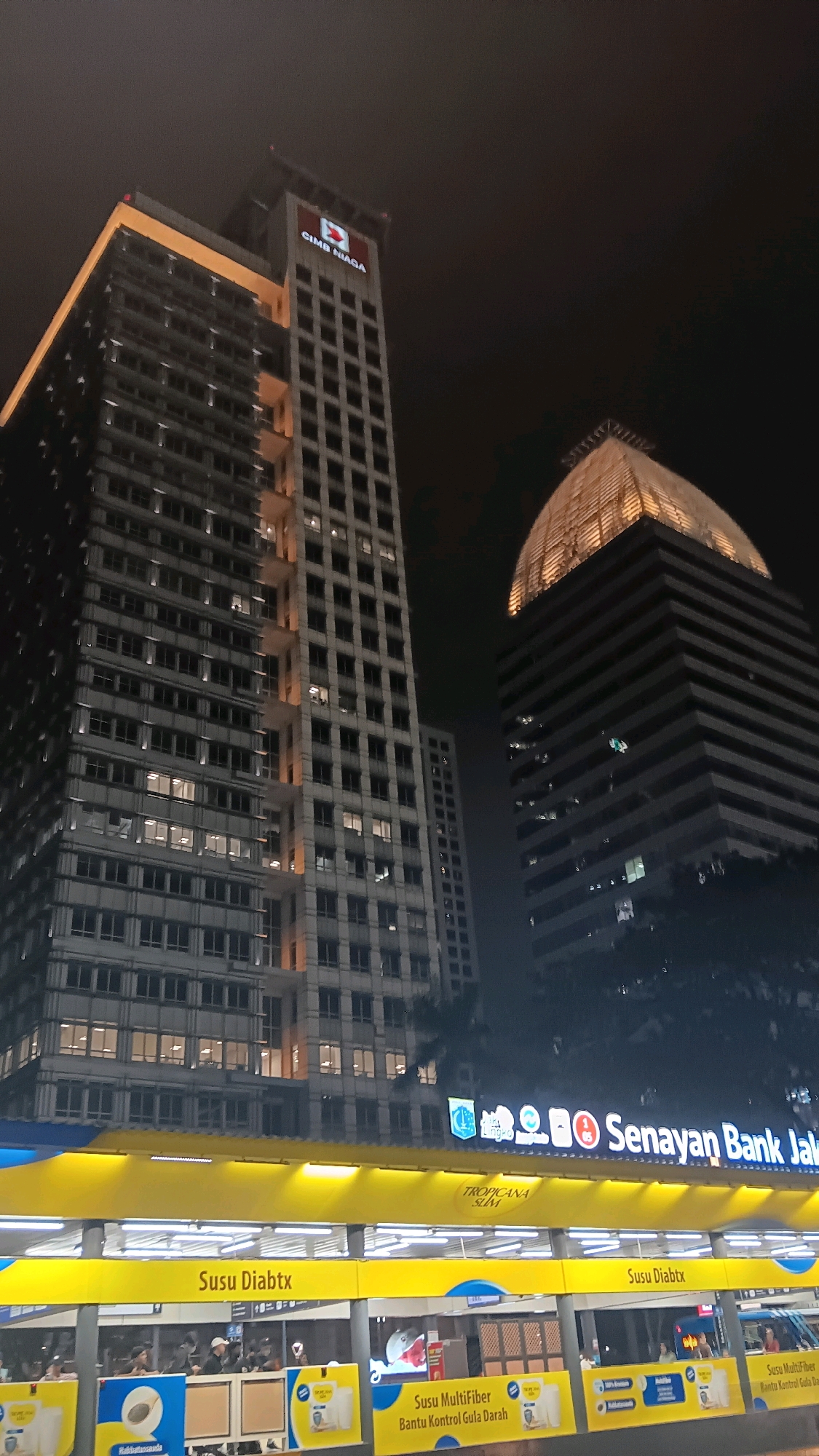 This is a nighttime, ground-level, wide-angle, vertical shot of a street scene in Central Jakarta, Indonesia, featuring a prominent bank building and a bus stop.
The dominant element is a tall, modern skyscraper with a grid of windows, illuminated from within, suggesting activity inside. The building's facade is a light color, possibly concrete or stone, with darker window frames. The top of the building features a large, illuminated logo with the text "CIMB NIAGA" in red and white. Another, shorter building with a distinct, illuminated, curved roof structure stands to the right of the main skyscraper.
In the foreground, a brightly lit yellow structure, likely a bus stop or transit station, spans the width of the image. This structure displays various advertisements, including one that reads "TROPICANA SLIM" and another with "Susu Diabtx". Below this, another level is visible with several people standing or walking, some appearing to be waiting for transportation. There are also various product advertisements on this lower level, featuring images of milk packaging and text in Indonesian, such as "Susu MultiFiber Bantu Kontrol Gula Darah" (MultiFiber Milk Helps Control Blood Sugar).
The sky is dark, indicating nighttime, and the overall lighting of the scene is provided by the building lights and the illuminated signage. The environment appears urban, with dense construction evident from the buildings. The overall impression is of a busy, well-lit commercial district at night.