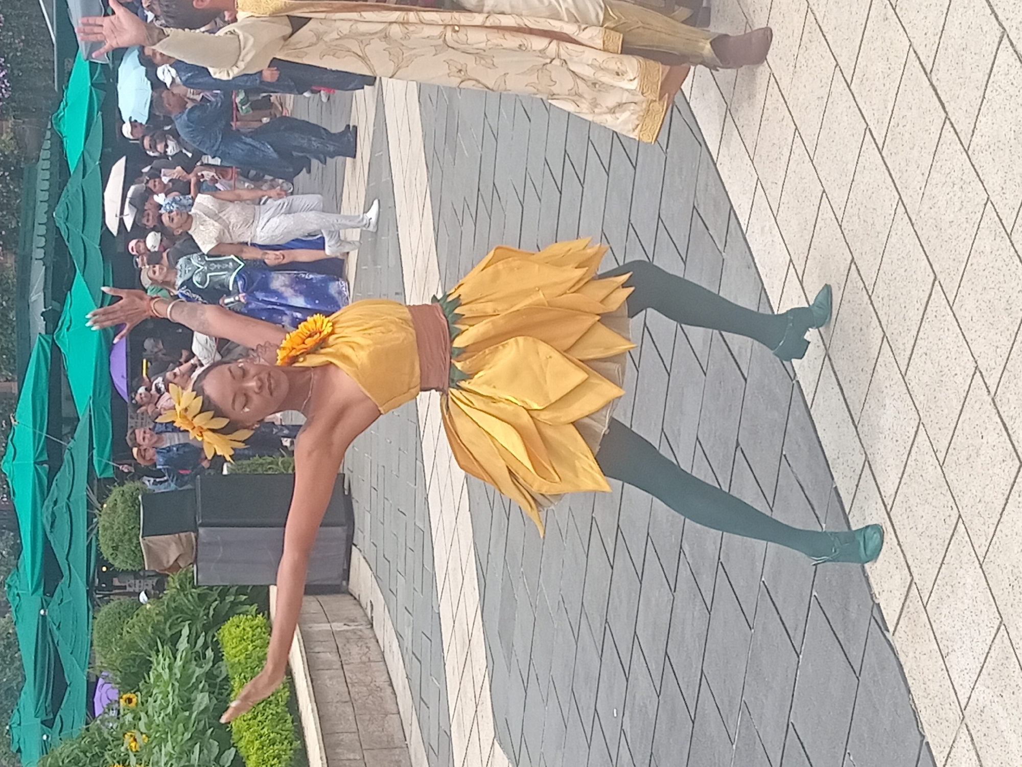 A woman in a yellow sunflower costume is dancing outdoors. She is wearing a yellow top with a flower accessory in her hair, a yellow skirt with petal-like detailing, and teal tights with teal heels. She is positioned in the foreground, with her arms outstretched. In the background, there are several people in costumes, some holding umbrellas, and trees with green canopies. The ground is paved with grey and white square tiles. The scene appears to be taking place at an outdoor event or festival. This event is likely at Sun World Ba Na Hills in Da Nang, Vietnam, given the stylized costumes and the presence of themed attractions.