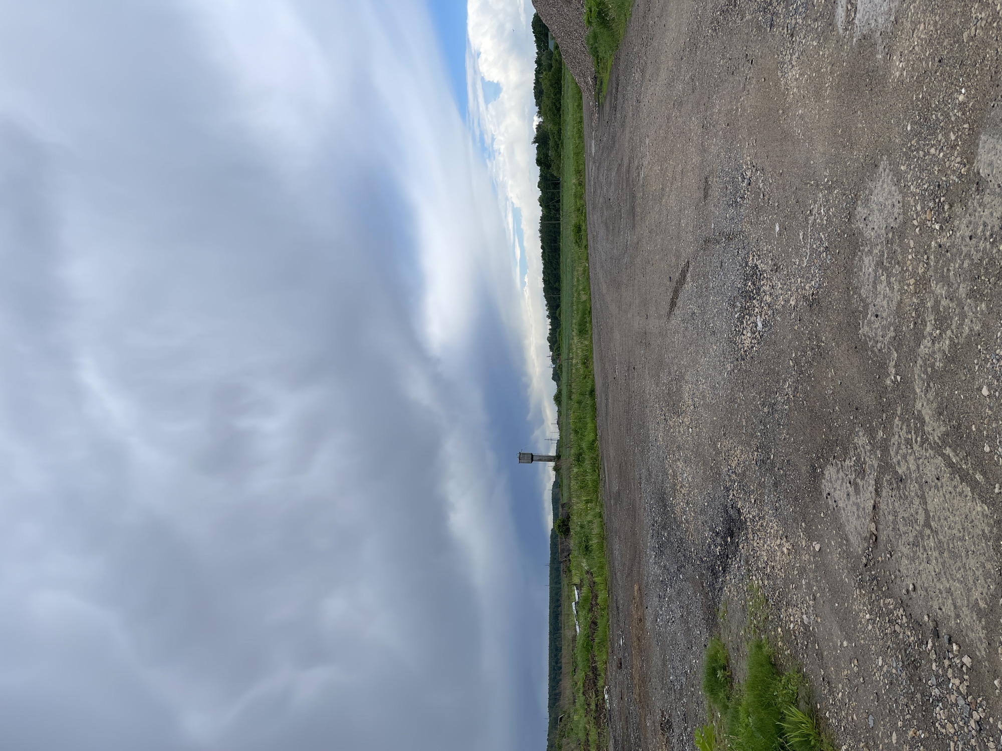 The image depicts a rural scene in Киясово, Russia. A dirt road stretches along the right side of the frame, transitioning to a green field with a line of trees in the center. A water tower rises above the horizon. The sky is dominated by dark, stormy clouds on the left, contrasting with patches of blue sky. The ground near the road is littered with small rocks and gravel.