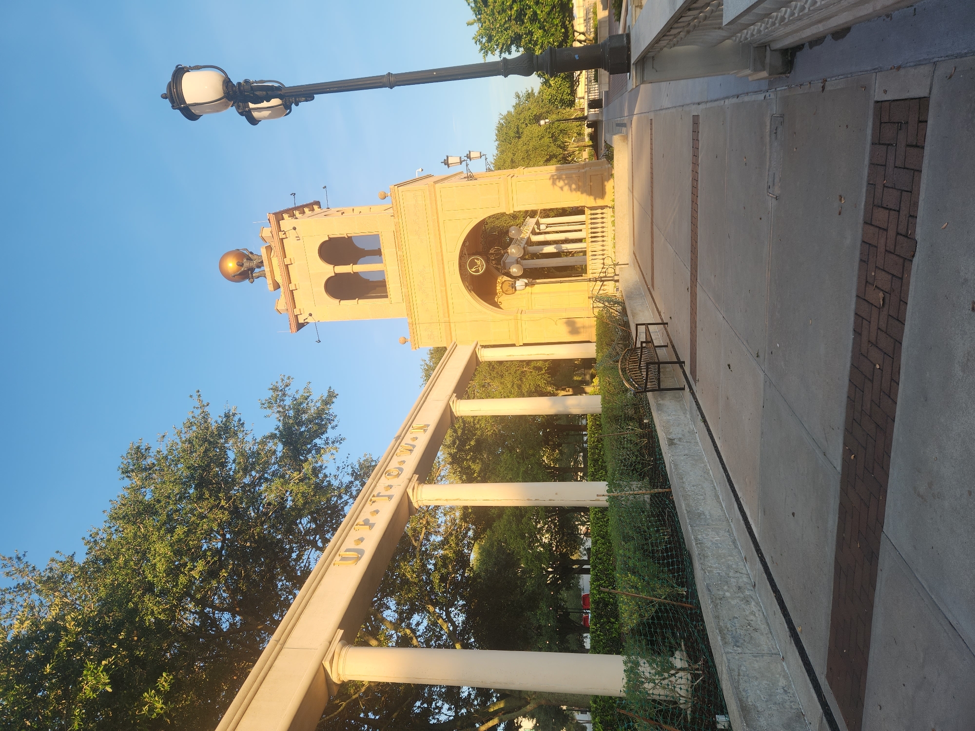 The image depicts a sunny outdoor scene in Altamonte Springs, Florida, United States of America. In the foreground, a paved walkway with decorative brick inlays leads towards a tan-colored tower. The tower features arched openings, decorative carvings, and a golden globe topped with figures. To the left of the walkway, a low wall borders a manicured garden with green bushes. Arching over the walkway, a pale structure with columns displays the golden letters "U P T O W N". A black park bench is visible on the left side of the walkway, and a tall black lamppost with multiple lights stands to the right. Lush green trees and a clear blue sky are visible in the background.