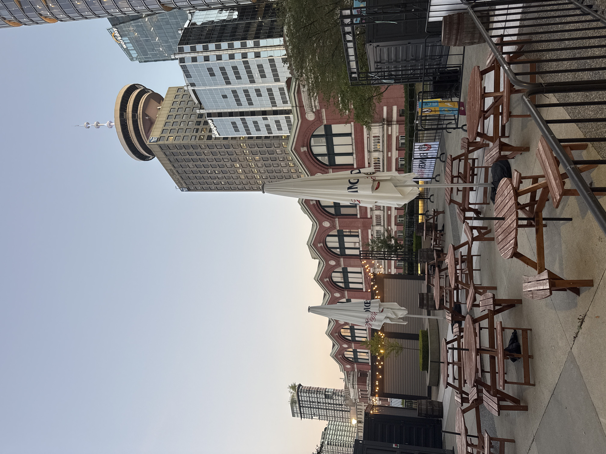 This image captures an outdoor seating area with multiple wooden picnic tables and benches, some with umbrellas. In the background, a red brick building with arched windows and decorative trim stands prominently, adorned with string lights. Beyond this, a modern cityscape unfolds, featuring skyscrapers, including the distinctive Harbour Centre with its observation deck and spire. The sky is a clear, pale blue, suggesting it's late afternoon or early evening. The scene appears to be set in an urban plaza or patio, likely a public space or part of a restaurant or event venue, within Vancouver, Canada. There are no people visible in the frame.