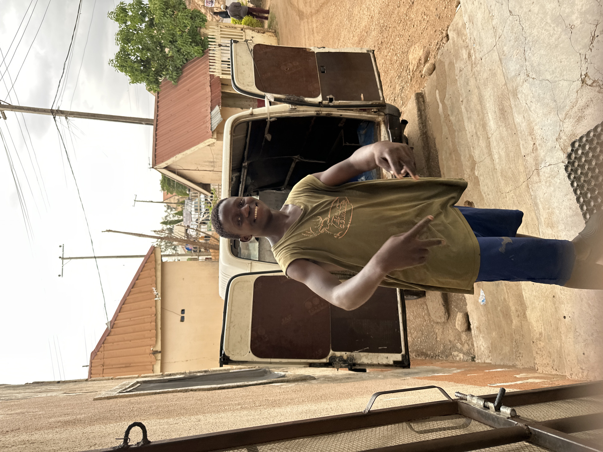 A smiling young man stands in front of a white van with open back doors on a dirt street in Jos, Nigeria. The man wears a green t-shirt and blue pants and is making a hand gesture. Buildings with brown roofs are visible behind the van. Power lines extend across the sky. A wire mesh barrier is in the foreground. Other people are visible in the background, further down the street.