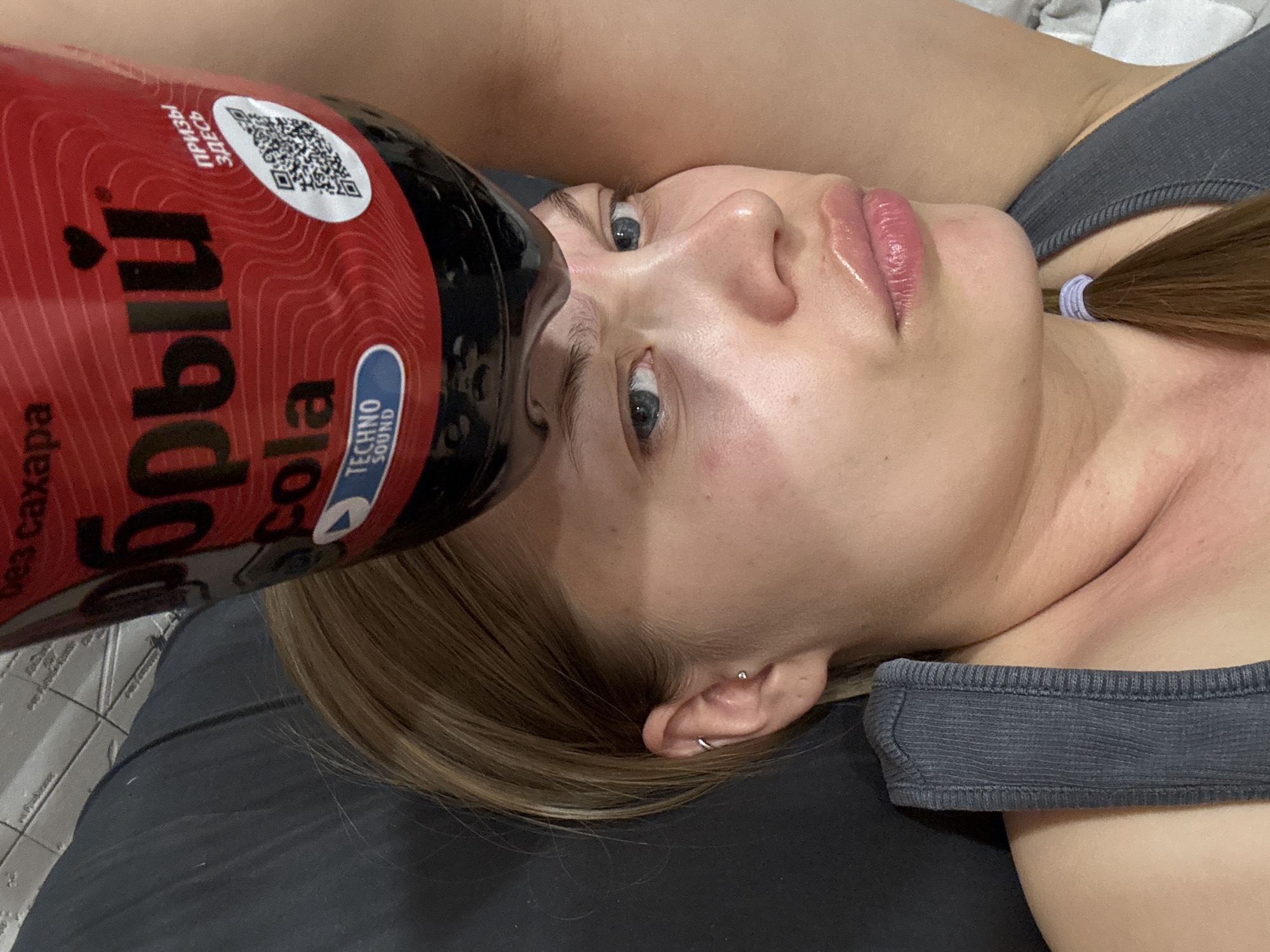 A woman is lying down, holding a bottle of Dobry Cola, a Russian cola brand, close to her face. The bottle is red with black lettering and a white QR code sticker. The label also includes the words "TECHNO SOUND" in English. The woman has blonde hair, fair skin, and blue eyes. She is wearing a gray tank top. The background includes a gray pillow or cushion. The city of Chaikovsky, Russia is not evident in the image.