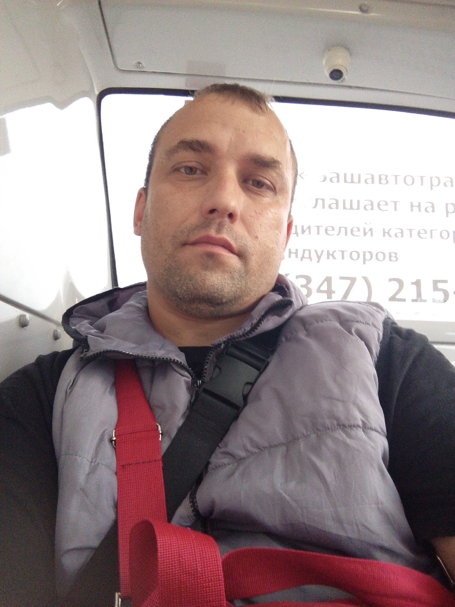 A man is taking a selfie in what appears to be a vehicle. He has short brown hair and stubble, and is wearing a black t-shirt and a gray puffer vest over it. A red strap is visible across his chest. Behind him, there is a window with text on it. The text is in Russian and appears to be an advertisement or informational sign. Some visible words include "вашавтотра", "лашает на р", "дителей категор", and "ндукторов", as well as a phone number "(347) 215-". A small security camera is mounted on the ceiling of the vehicle. The lighting suggests it is daytime.