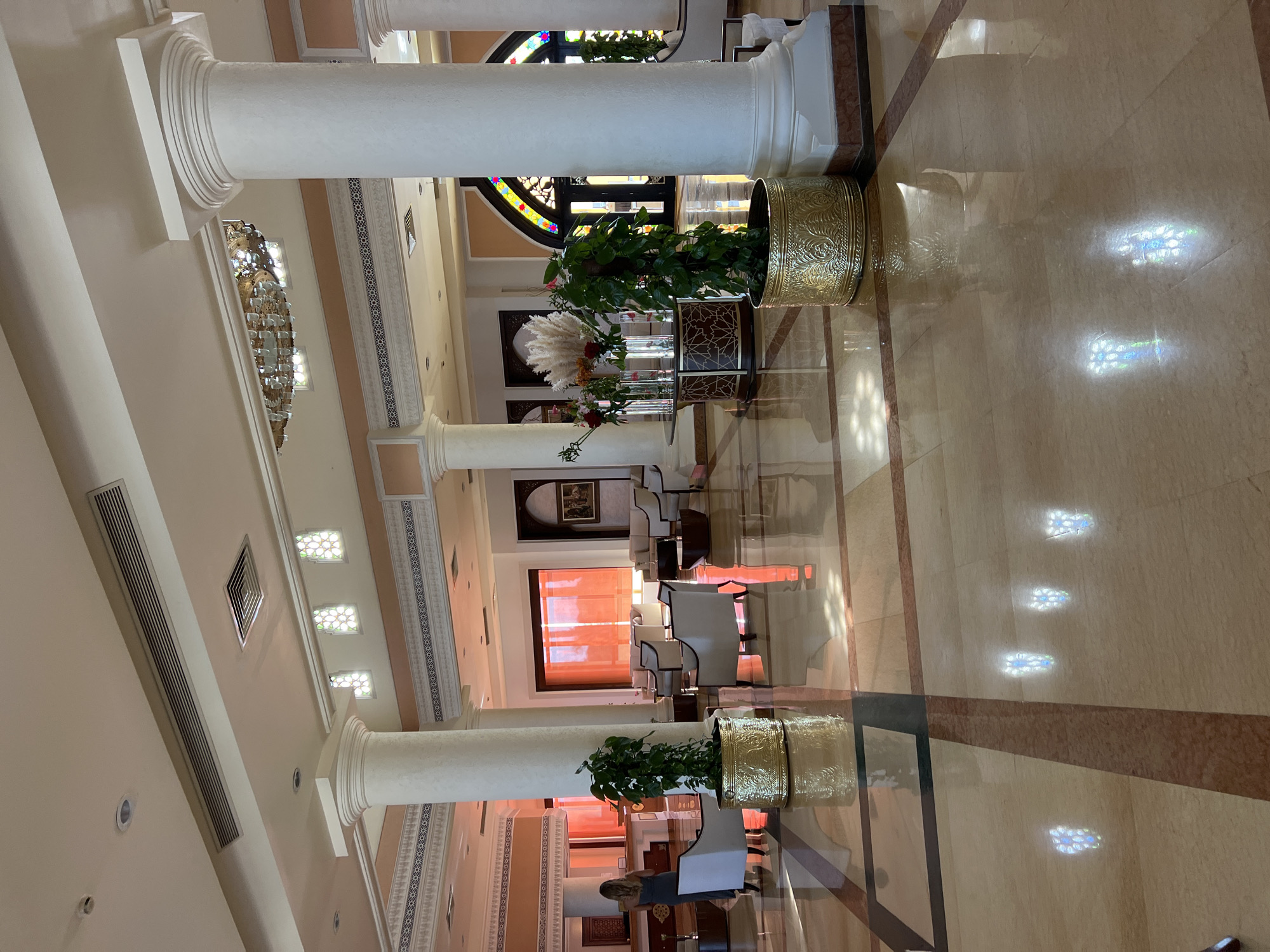 This is a vertical, slightly angled, full shot of a hotel lobby in Sharm El Sheikh, Egypt. The scene is well-lit and appears to be during the daytime, with sunlight casting reflections on the polished marble floor. The lobby features large, white, fluted columns that divide the space and create a sense of grandeur.
Several potted plants in ornate golden planters are strategically placed. One planter is particularly prominent in the foreground, containing a lush green plant. Further down the hall, another identical planter is visible. In the mid-ground, there's a display of dried reeds and flowers, adding a touch of natural decor.
The setting includes seating areas with white upholstered chairs arranged around low tables. A large window with intricate stained-glass elements and a dark frame is visible in the background, letting in diffused light. The walls are a neutral, warm tone, decorated with patterned borders and artwork. The ceiling has recessed lighting and air vents.
A woman is partially visible in the lower right portion of the image, facing away from the camera and appearing to be interacting with a reception desk. The overall atmosphere of the lobby is elegant and inviting, suggesting a high-end establishment. The light filtering through the stained glass creates colorful, patterned reflections on the floor, adding a dynamic visual element to the scene.