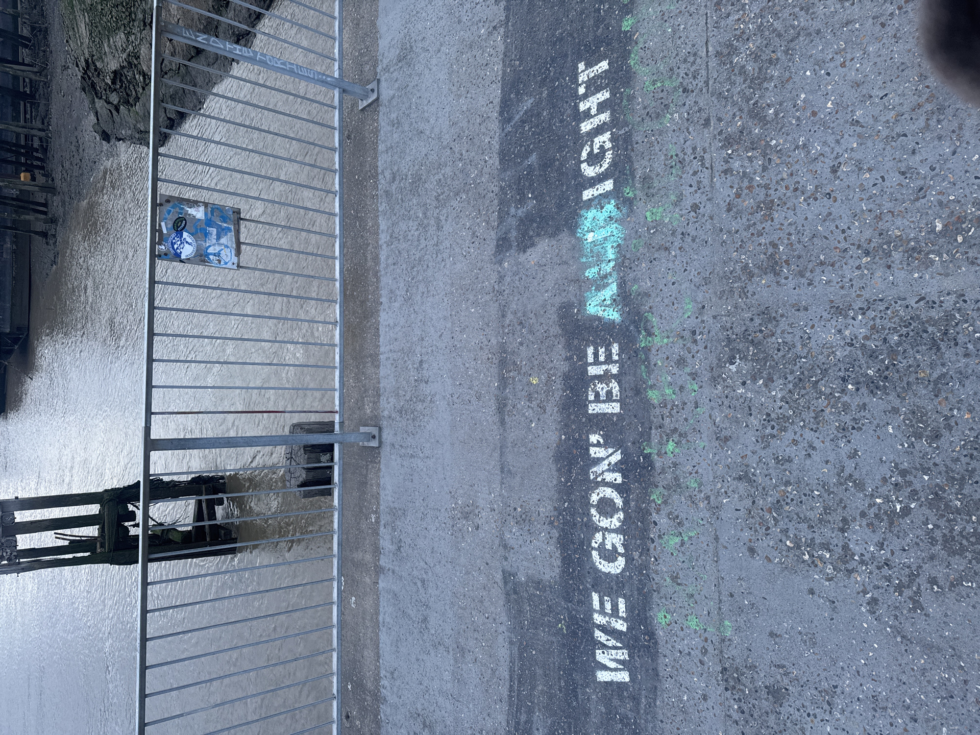 A vertical, outdoor shot depicts a concrete surface with the words "WE GON' BE ALRIGHT" painted in white with traces of green. A metal fence runs along the left side of the frame, separating the concrete from what appears to be a body of water with wooden structures submerged in it. A sign with stickers is attached to the fence. The scene is located on the bank of the River Thames in the city of London, United Kingdom.