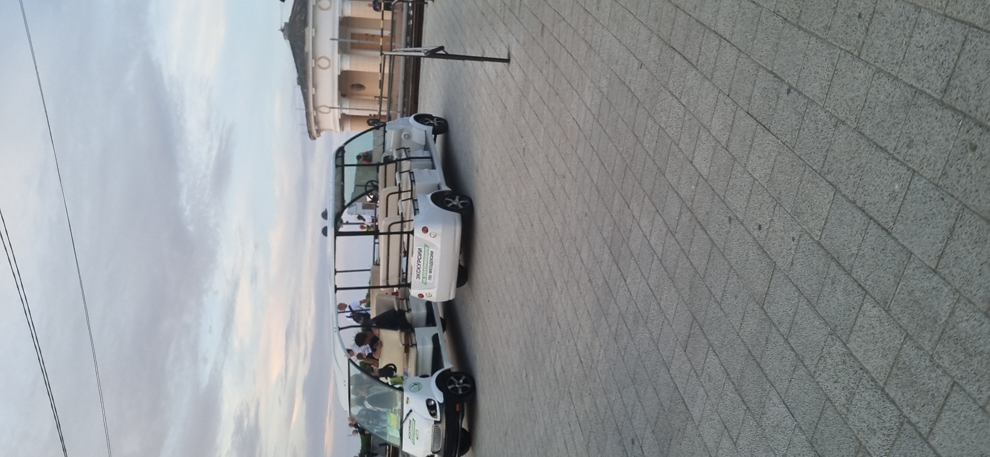 The image shows two white open-air electric vehicles parked on a paved walkway. The vehicle on the left has passengers inside. The vehicle on the right has text on its rear that reads "ЭКСКУРСИИ ПО ФЕОДОСИИ" which translates to "EXCURSIONS IN FEODOSIA". In the background, a circular building with columns is visible. The sky is overcast. The paved walkway is made of rectangular grey tiles.