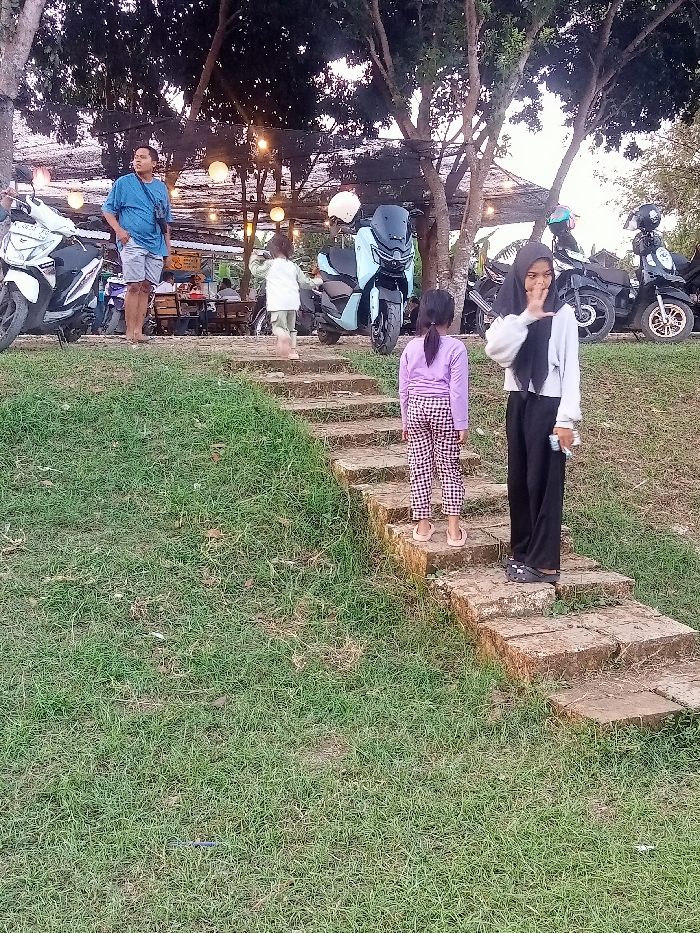 The scene is a grassy area with a set of stone steps. There are four people present, including a man in a blue shirt and a woman wearing a hijab. Two young girls are standing on the steps. There are several motorbikes parked nearby, including a light blue scooter and a black motorbike. The scene is located in Jakenan, Indonesia, at the coordinates -6.758 latitude and 111.075 longitude. There is a wooden structure in the background with string lights hanging from it. The scene is lit by natural sunlight and the artificial lights. The grassy area is covered with lush green grass. There are trees surrounding the grassy area and the wooden structure.