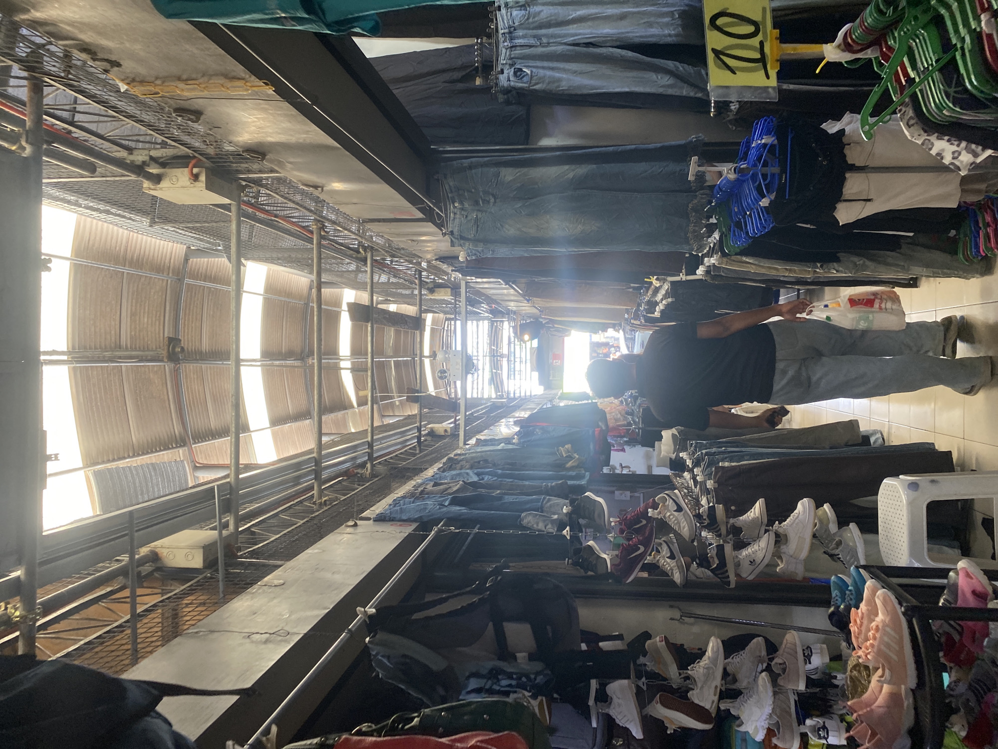 The image shows an active outdoor or semi-outdoor market stall in Davao City, Philippines, under bright sunlight, likely during the day. The scene is oriented vertically, but depicts a long, narrow pathway lined with merchandise on both sides, and an elaborate, exposed metal framework overhead supporting a corrugated roof.
In the mid-ground, a person is seen from behind, wearing a dark t-shirt and light pants, holding a white plastic shopping bag. They appear to be browsing or shopping among the goods. Adjacent to them, various items of clothing, including jeans and other garments, are displayed on hangers and folded on racks. Numerous pairs of shoes, including sneakers in different styles and colors, are neatly arranged on multi-tiered stands. A white plastic stool is also visible near the merchandise.
Above the merchandise, a yellow sign with the number "20" written in black is clearly visible, likely indicating a price or an item count. The structure overhead features extensive scaffolding, metal beams, and wire mesh trays, through which sunlight streams, creating bright highlights and lens flares. The far end of the pathway fades into bright light, suggesting a long corridor or extensive market area. Electrical conduits and junction boxes are integrated into the overhead framework, indicating functional infrastructure within this open-air setting.