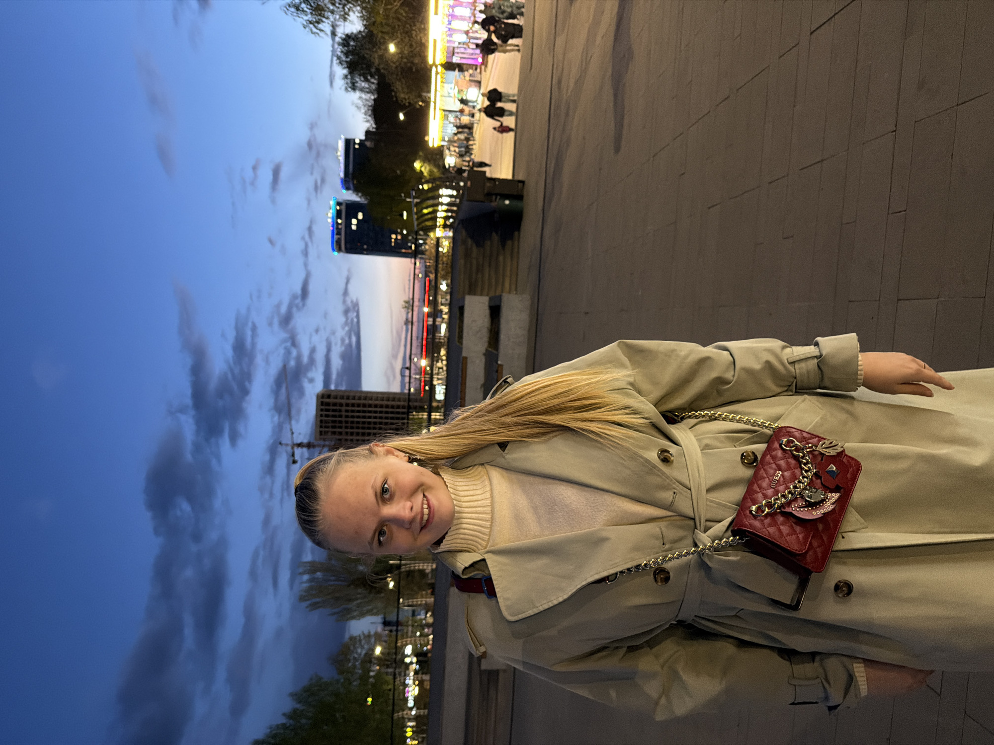 The image shows a woman standing outdoors in Izhevsk, Russia, at dusk. She is fair-skinned with blonde hair pulled back, wearing a beige trench coat over a light-colored sweater and carrying a small red quilted purse with a gold chain strap. The coat is belted at the waist. The background includes a paved walkway, a cityscape with buildings, streetlights, and a cloudy blue sky. Some people are walking along the walkway in the distance. The overall setting suggests an urban, possibly waterfront location.