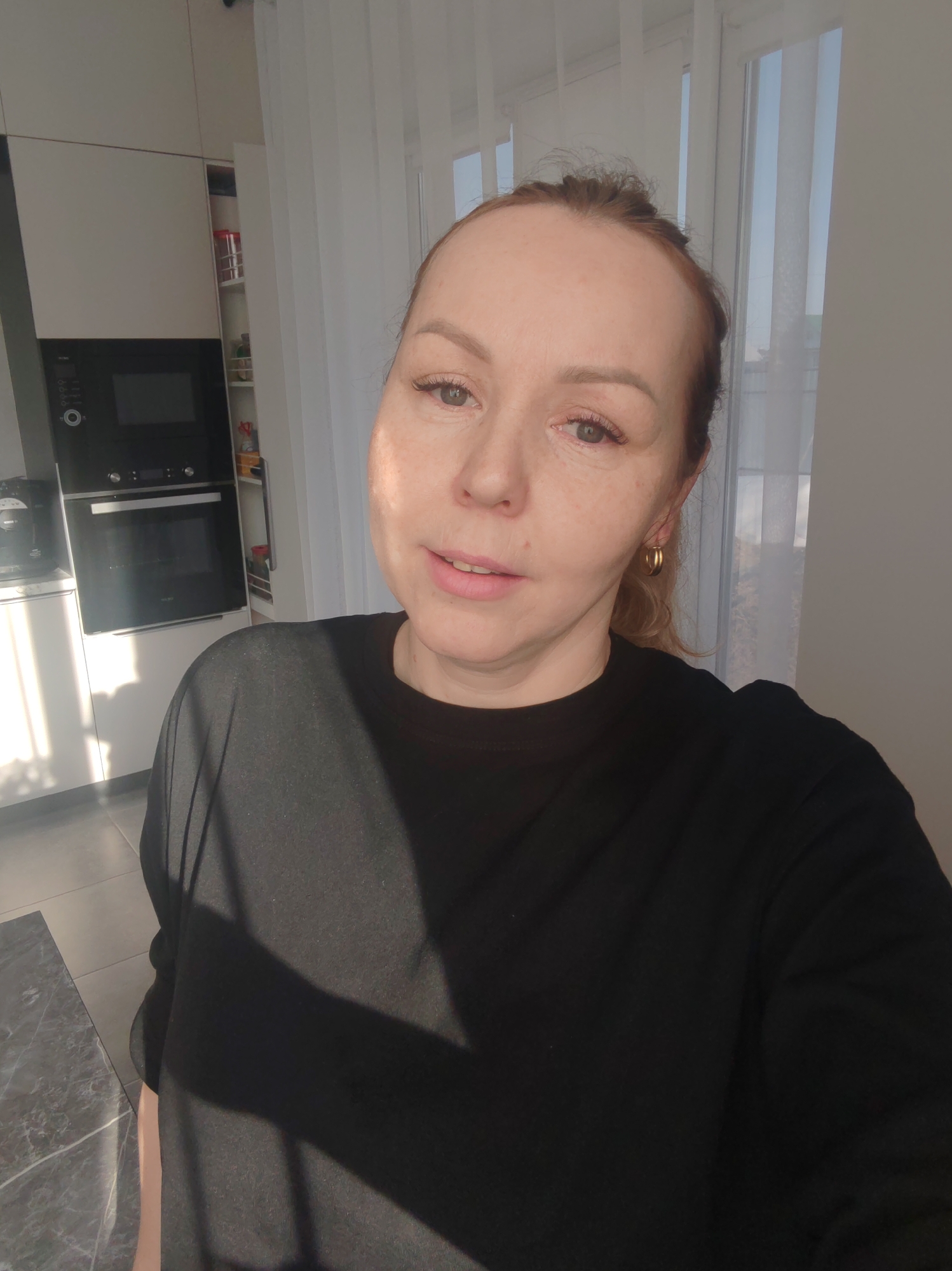 A Caucasian woman with light skin and auburn hair, casually styled, is taking a selfie indoors. She wears a black top and gold hoop earrings. The background suggests a modern kitchen with white cabinets and a built-in black oven. Sunlight streams in from a window covered with sheer white curtains. Based on the user's location, the image was taken in Chaikovsky, Russia.