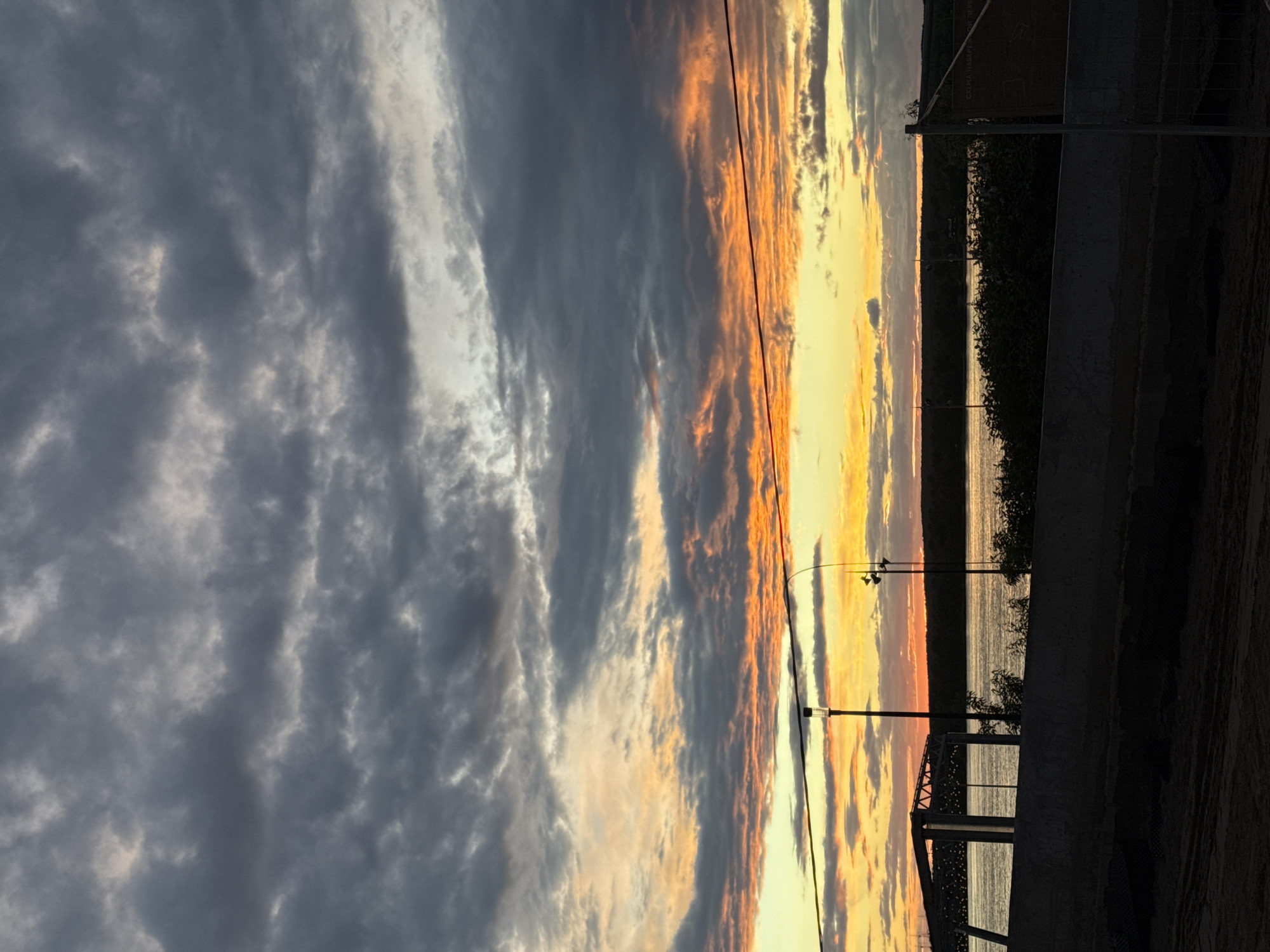 The image captures a sunset over the Kama River in Perm, Russia. The sky is filled with clouds, ranging from dark gray to shades of orange and yellow. The river reflects the colors of the sky. In the foreground, there's a structure, possibly a bridge or part of a dock. Vegetation is visible along the riverbank. Electrical lines stretch across the sky, adding to the urban landscape.