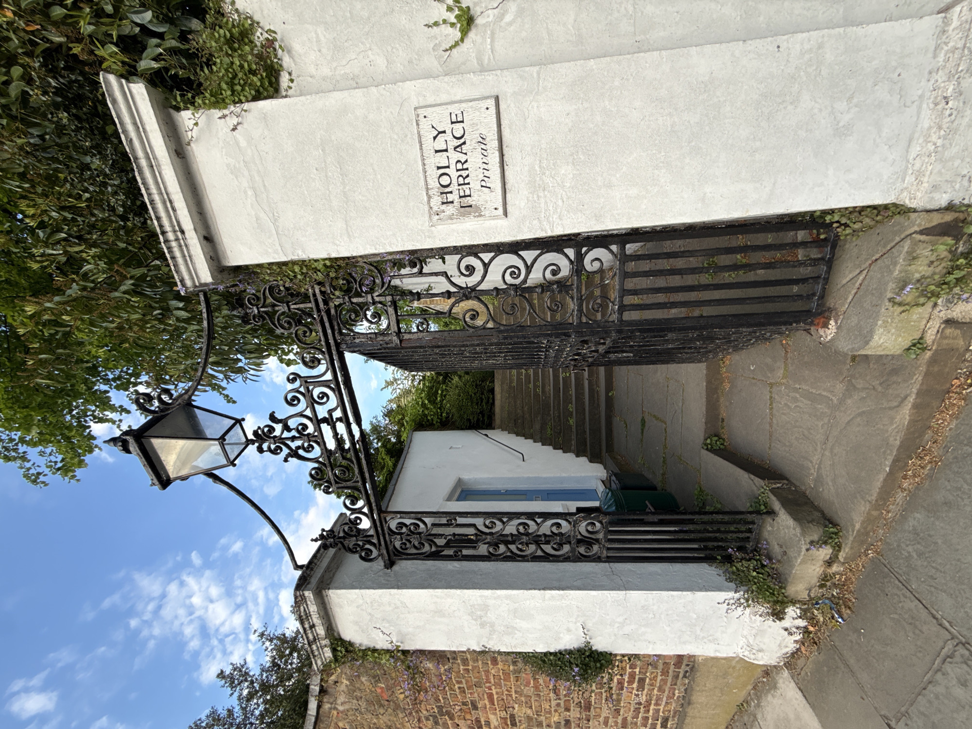 The image depicts an entrance to "Holly Terrace Private" in London, United Kingdom. The entrance features a white wall with a sign indicating the name of the terrace. A black wrought-iron gate and an ornate lamppost stand at the entrance. Beyond the gate, steps lead up to a white building with a blue door and window frames. The setting includes brickwork, paving stones, and some greenery, suggesting an urban environment. The sky is visible with scattered clouds.