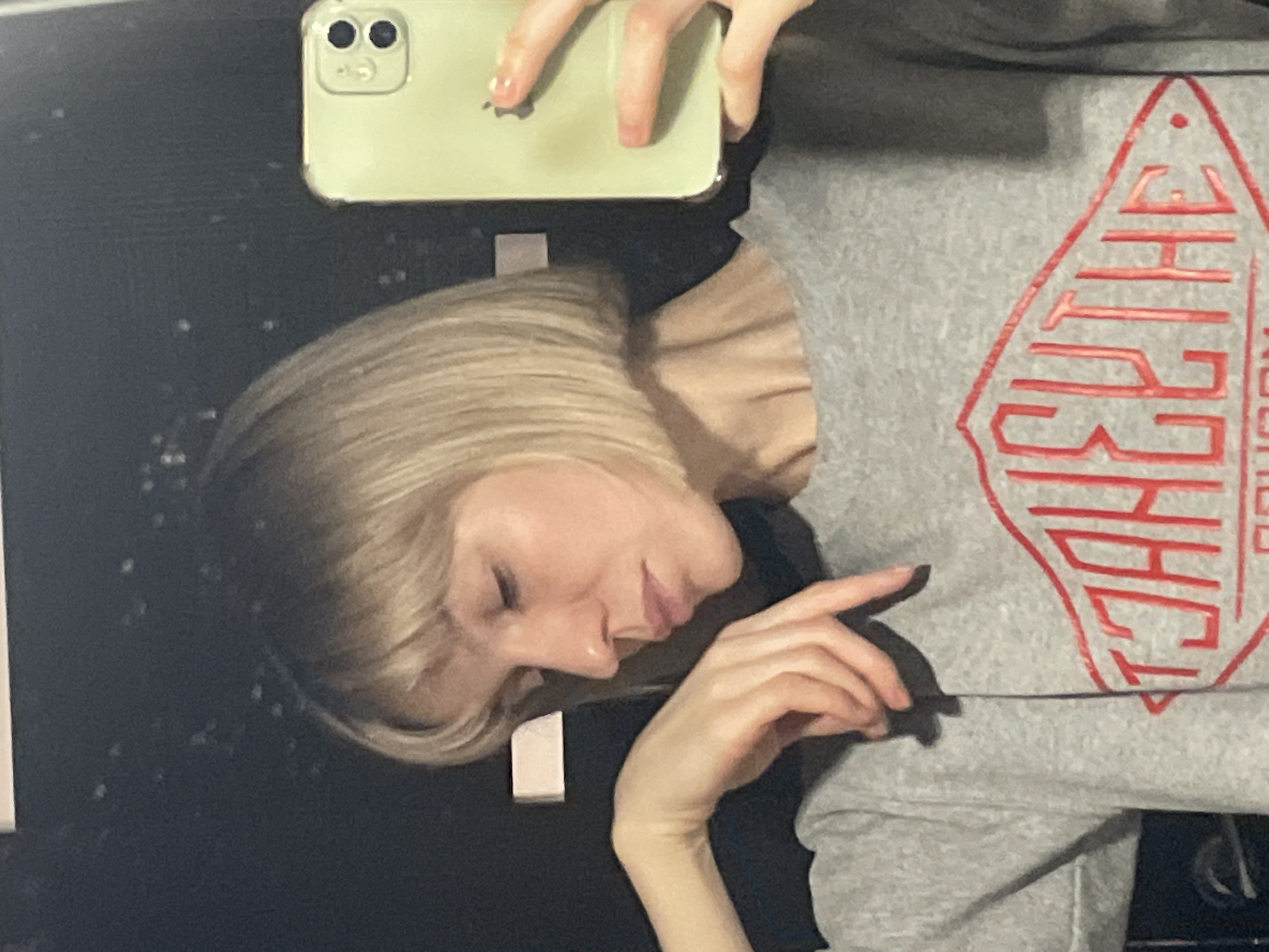 A person with blonde hair is holding a light green phone, possibly taking a selfie in front of a black surface. The person is wearing a grey sweatshirt with a red logo. The location is unknown.