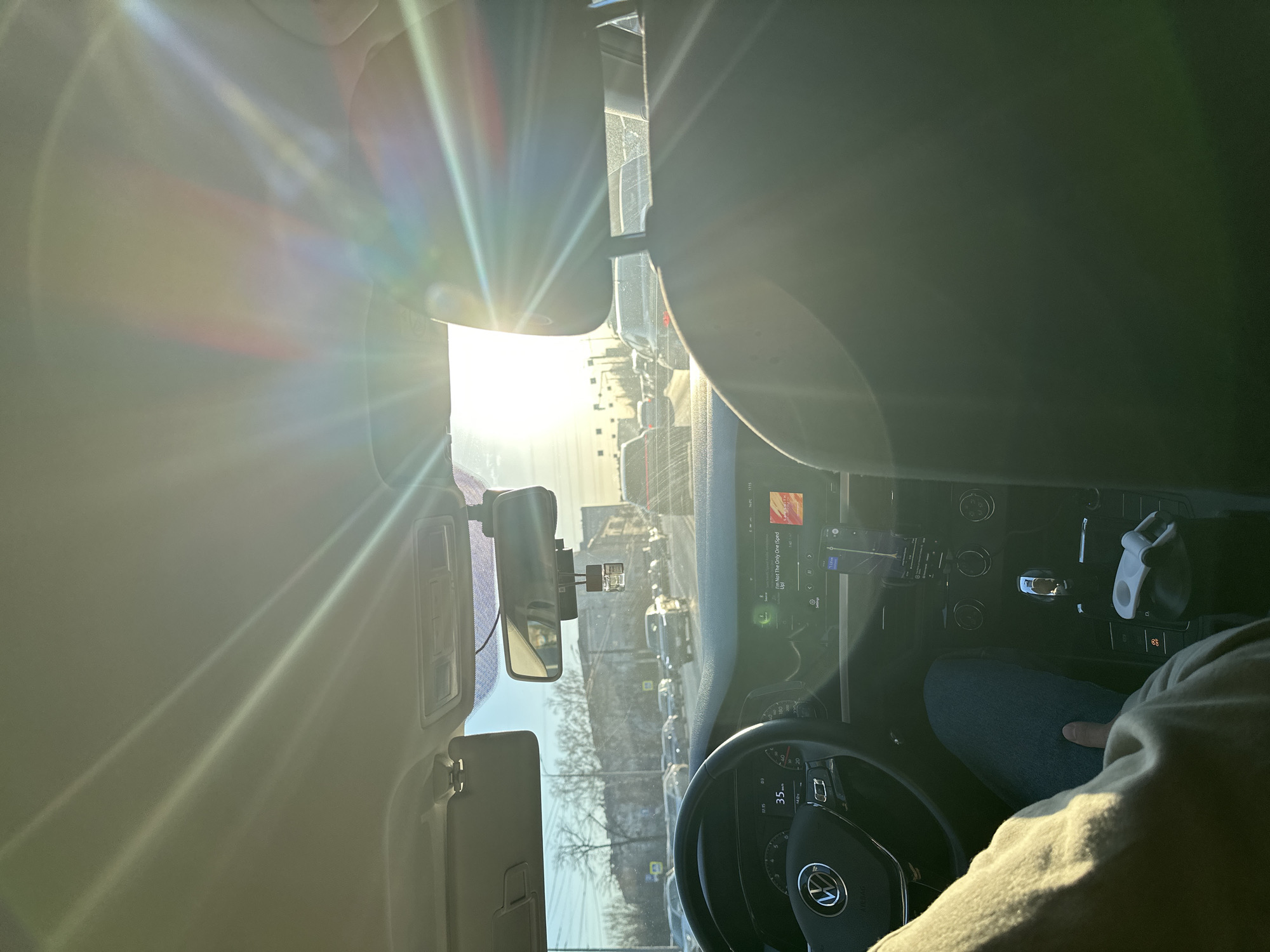 The image shows the interior of a car, likely a Volkswagen, in a stationary position amidst heavy traffic in Kazan, Russia. The sun shines brightly ahead, causing lens flare and obscuring the view. The dashboard is visible, with a digital display showing the speedometer reading 35 km/h. The steering wheel has a Volkswagen logo. To the left, there is a rearview mirror and sun visor. The driver, partially visible, is wearing jeans and a light-colored jacket. Outside, a street lined with trees, buildings, and numerous cars is visible. Road signs are also present. The overall environment indicates a busy city street during daylight hours.