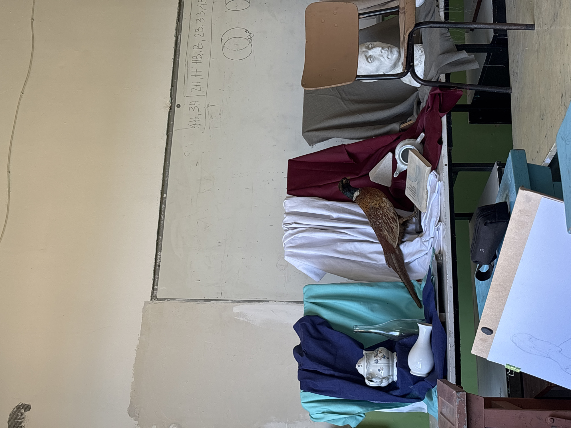 The image displays a still life arrangement within what appears to be an art classroom or studio in Troyan, Bulgaria. The original image is oriented 90 degrees counter-clockwise.
A whiteboard occupies the left side of the frame, showing handwritten text listing pencil lead hardness grades: "4H, 3H", "2H, H, HB, B, 2B, 3B, 4B". Below this, simple diagrams of cylindrical and circular shapes are drawn. The walls are a light, somewhat worn beige.
The central subject is a still life setup. A taxidermied pheasant with detailed brown, black, and reddish feathers, and a dark head, is prominently positioned. It rests on white fabric, with other draped cloths in maroon, teal, and dark blue providing a backdrop and foundation. Other items in the still life include a white ceramic head sculpture, a small white teapot adorned with a floral pattern, a plain white vase, a clear glass bottle, a small white cup or bowl, and an open book.
To the right, parts of a wooden chair with a metal frame and black metal stands (likely easels) are visible. A drawing board, clipped to an easel, shows a partial pencil sketch, reinforcing the art studio context. A black pouch or bag is also seen. The scene implies an ongoing or recently concluded art drawing session, though no people are present. The lighting is uniform, and no specific time of day or weather is discernible.