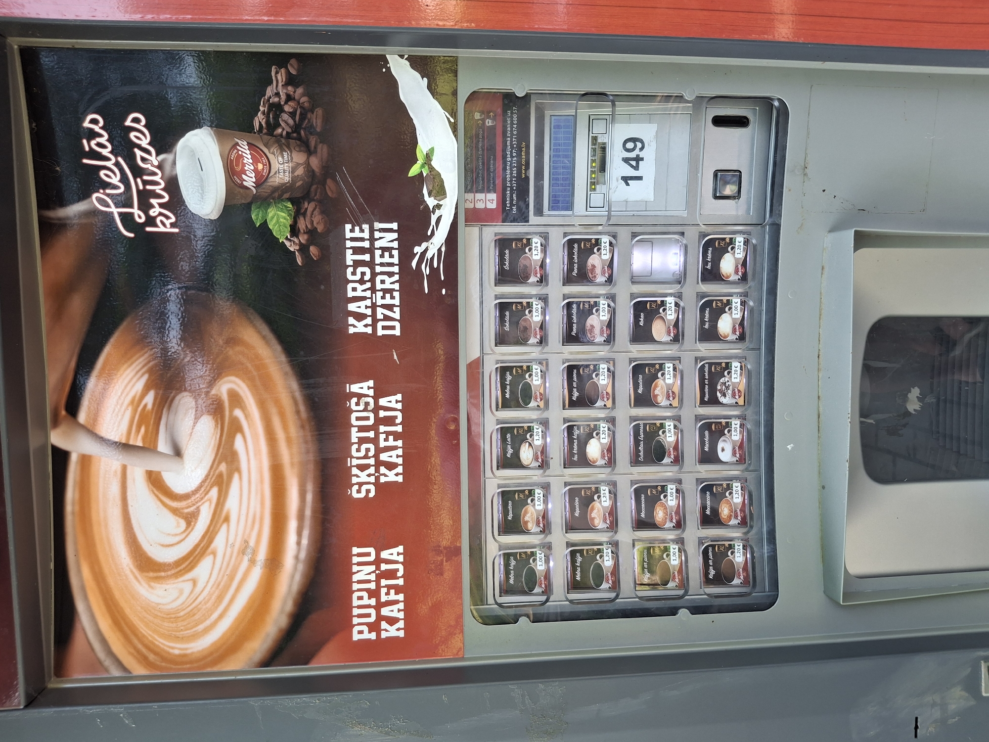 The image shows a Merill coffee vending machine located in Lēdmane, Latvia. The machine is in a public area, likely outdoors, with a red-colored wall visible on the right side. The top portion of the machine features an advertisement with an image of latte art and a cup of coffee. The advertisement includes text in Latvian: "Lielās krūzes" (large cups), "Pupinu kafija" (bean coffee), "Šķīstošā kafija" (instant coffee), and "Karstie dzērieni" (hot drinks). Below the advertisement is a display of various coffee and hot drink options with their prices, indicated in Euros. To the right of the drink selections, there is a control panel with a digital display showing "149", a coin slot, and buttons. The machine appears to be in working order.