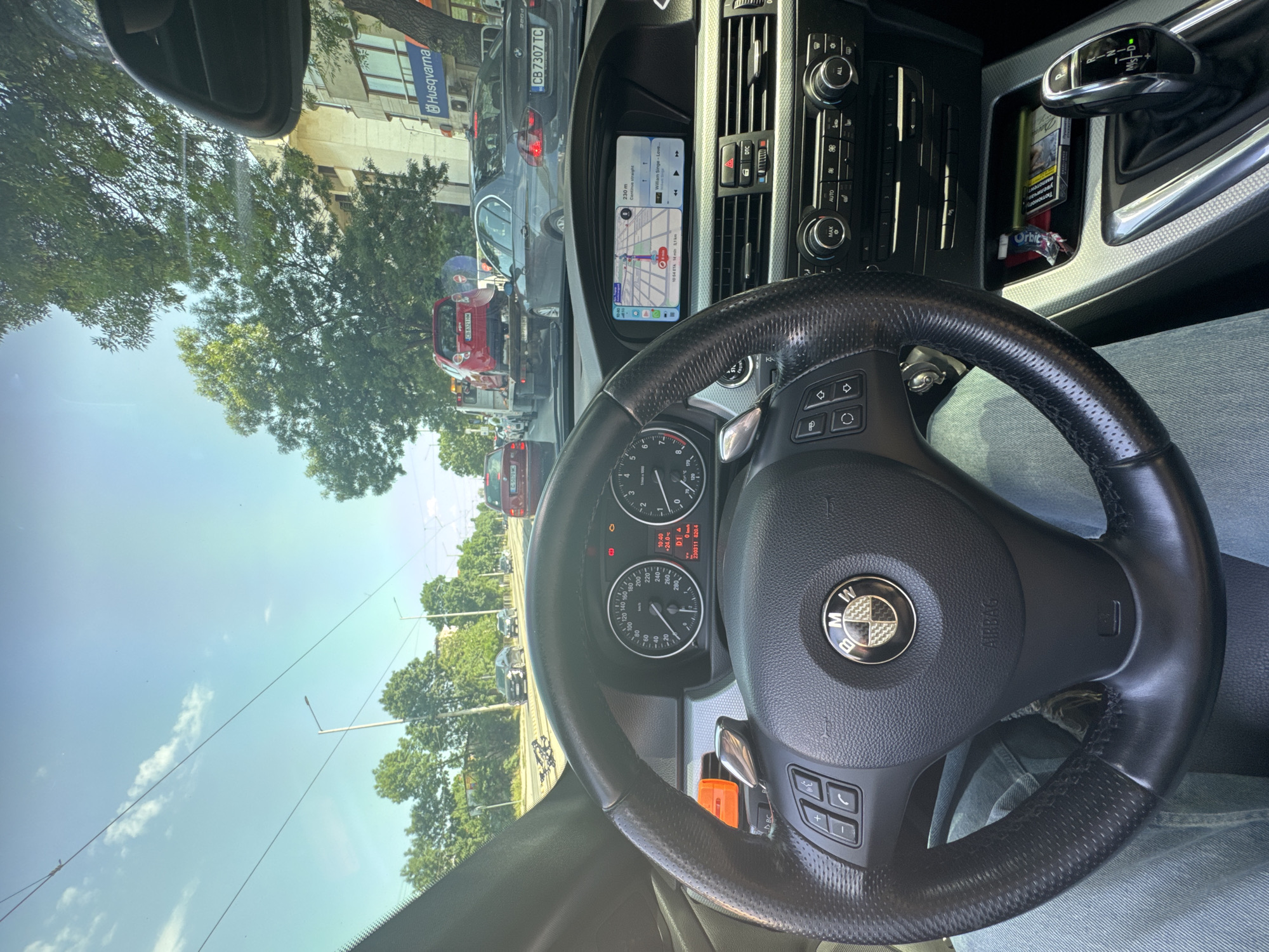 A view from inside a BMW car, showing the steering wheel, dashboard, and a glimpse of the road outside. The navigation system displays a route. Outside, there's traffic, including a red car being towed. A "Husqvarna" sign is visible on a building across the street. The dashboard clock reads 10:40 with a temperature of +24.0 degrees Celsius. The license plate "CB 7307 TC" is visible on a vehicle, as well as "5070 MC" and "086321HM" on others. Cigarettes and gum are in the console next to the gear stick.