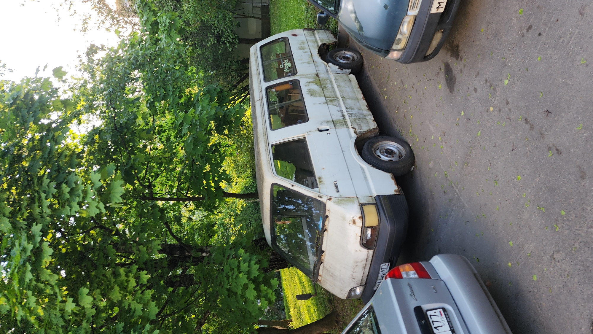 An outdoor scene shows a dilapidated white van parked on a paved surface, possibly a parking area or side street. The van exhibits significant signs of rust and wear, suggesting long-term neglect or abandonment. It is parked between two other vehicles, one green and the other silver. A license plate visible on the silver car indicates that the scene is in Moscow, Russia (license plate beginning with "77" or "177" indicates Moscow). Lush green trees surround the parking area, with leaves scattered on the pavement. The overall environment appears urban and potentially residential. No people are visible in the frame. The light indicates a daytime scene.
