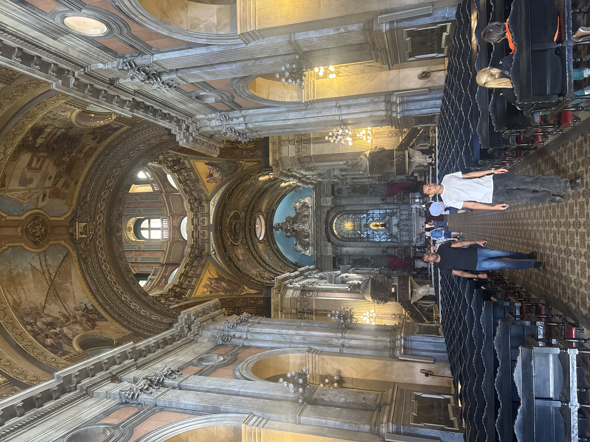 The image captures the interior of the Metropolitan Cathedral of Rio de Janeiro, Brazil, a vast and ornate religious structure. In the foreground, two men walk down the central aisle, which is flanked by rows of dark wooden pews. The man on the left wears a black t-shirt and blue jeans, while the man on the right is in a white t-shirt and dark pants. Further down the aisle, several other individuals are visible, including a woman with blonde hair seated on a pew and two people in the distance walking towards the altar.
The cathedral's architecture is grand, with soaring vaulted ceilings adorned with intricate frescoes and gilded detailing. Large marble columns support the structure, and ornate chandeliers hang from the ceiling, casting a warm glow. The altar area, visible in the background, is richly decorated with sculptures and stained-glass windows. The overall impression is one of immense scale, historical significance, and a serene atmosphere suitable for worship and contemplation.