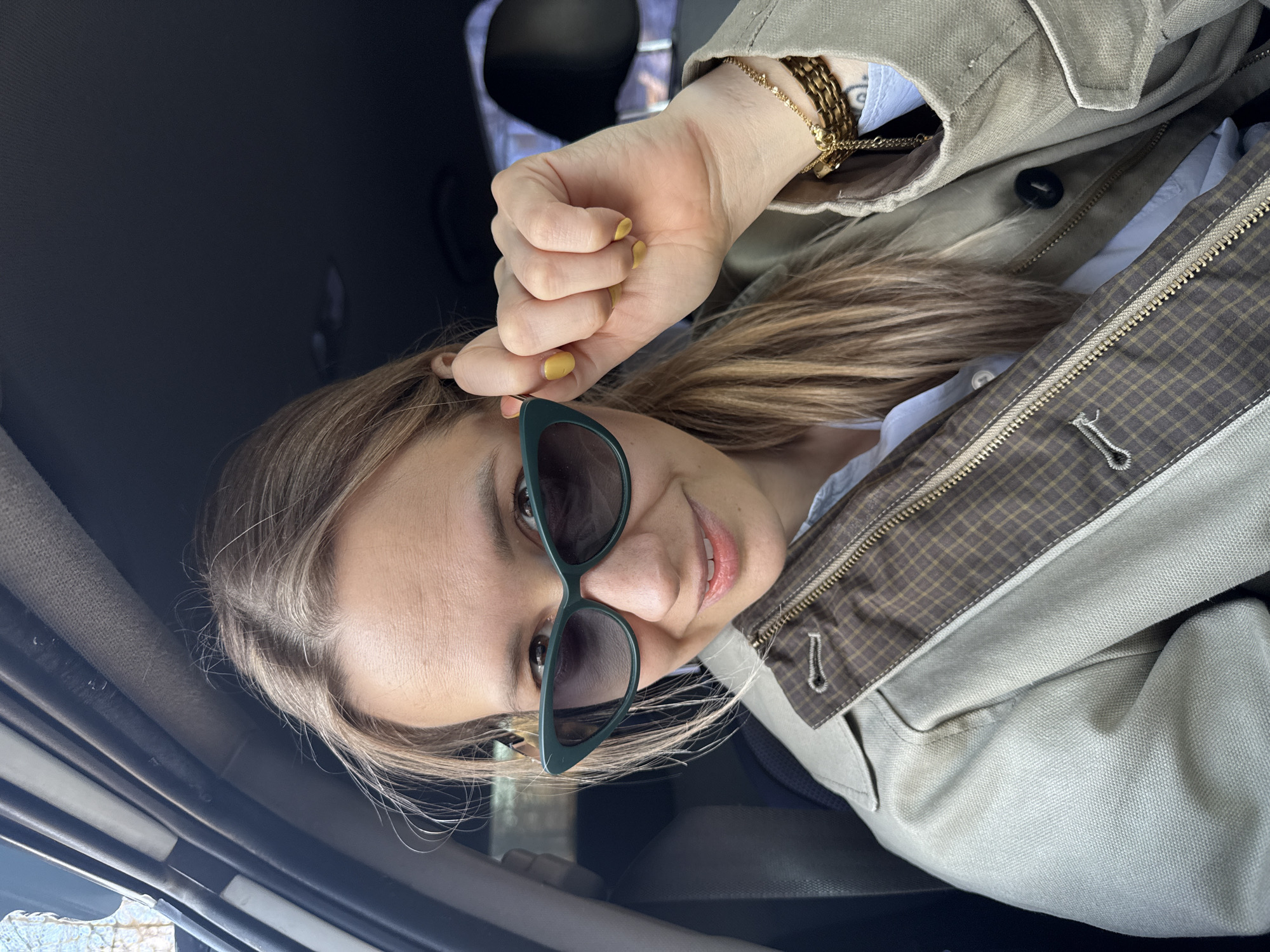 The image depicts a woman inside a vehicle, possibly a car. She is wearing sunglasses with a green frame and a beige jacket over a light blue shirt. Her hair is blonde, and her nails are painted yellow. She is holding the side of her sunglasses with one hand, and she is smiling at the camera. The interior of the vehicle is dark. The location is not identifiable as Chaikovsky, Russia.